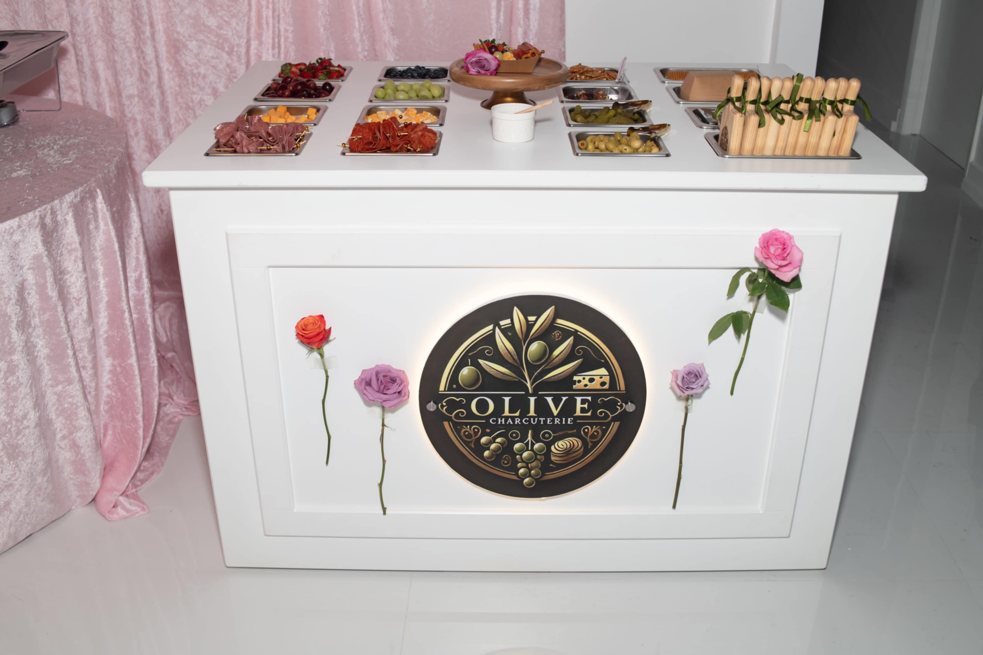 A white charcuterie table is adorned with a variety of food items in trays, featuring a decorative logo and fresh flowers on the front.
