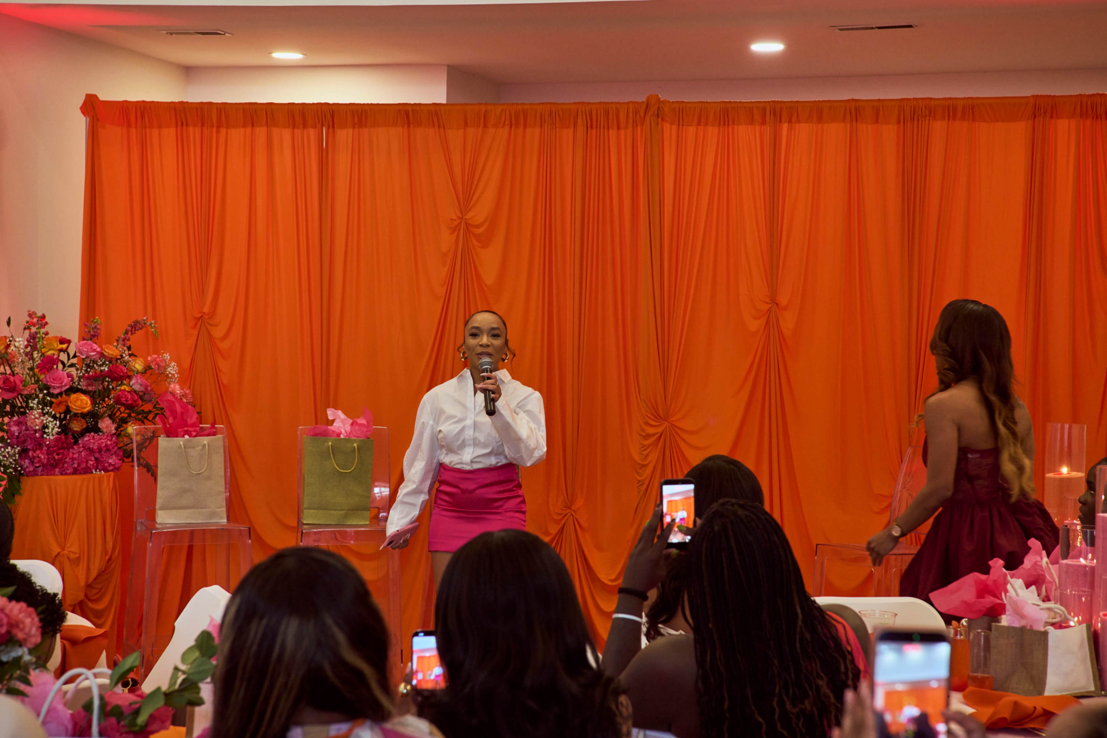 A woman stands on stage speaking into a microphone with gift bags and a vibrant orange backdrop in a decorated event space.