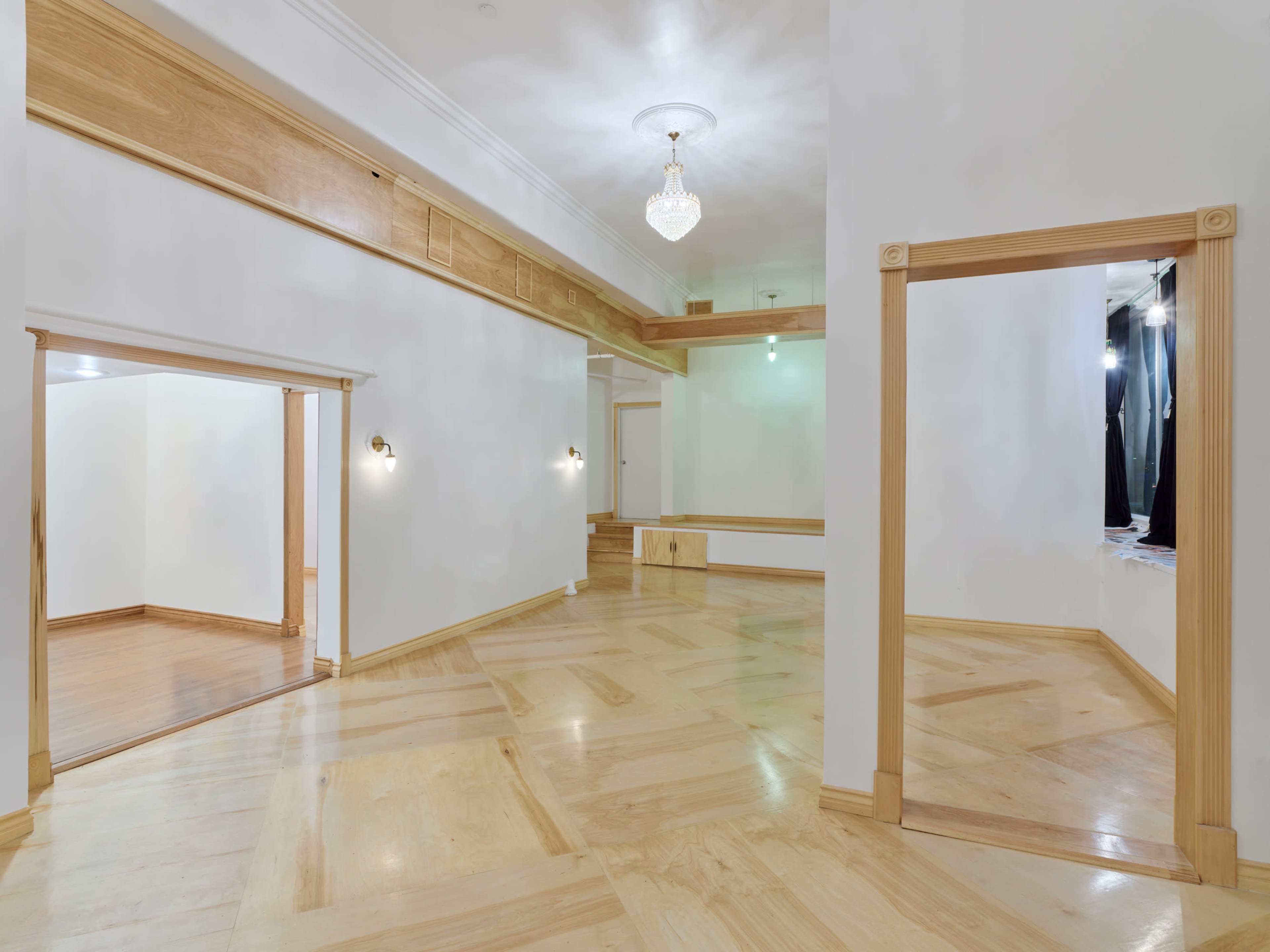 The image shows a spacious, well-lit interior room with wooden flooring and minimalistic design elements, featuring two open doorways.