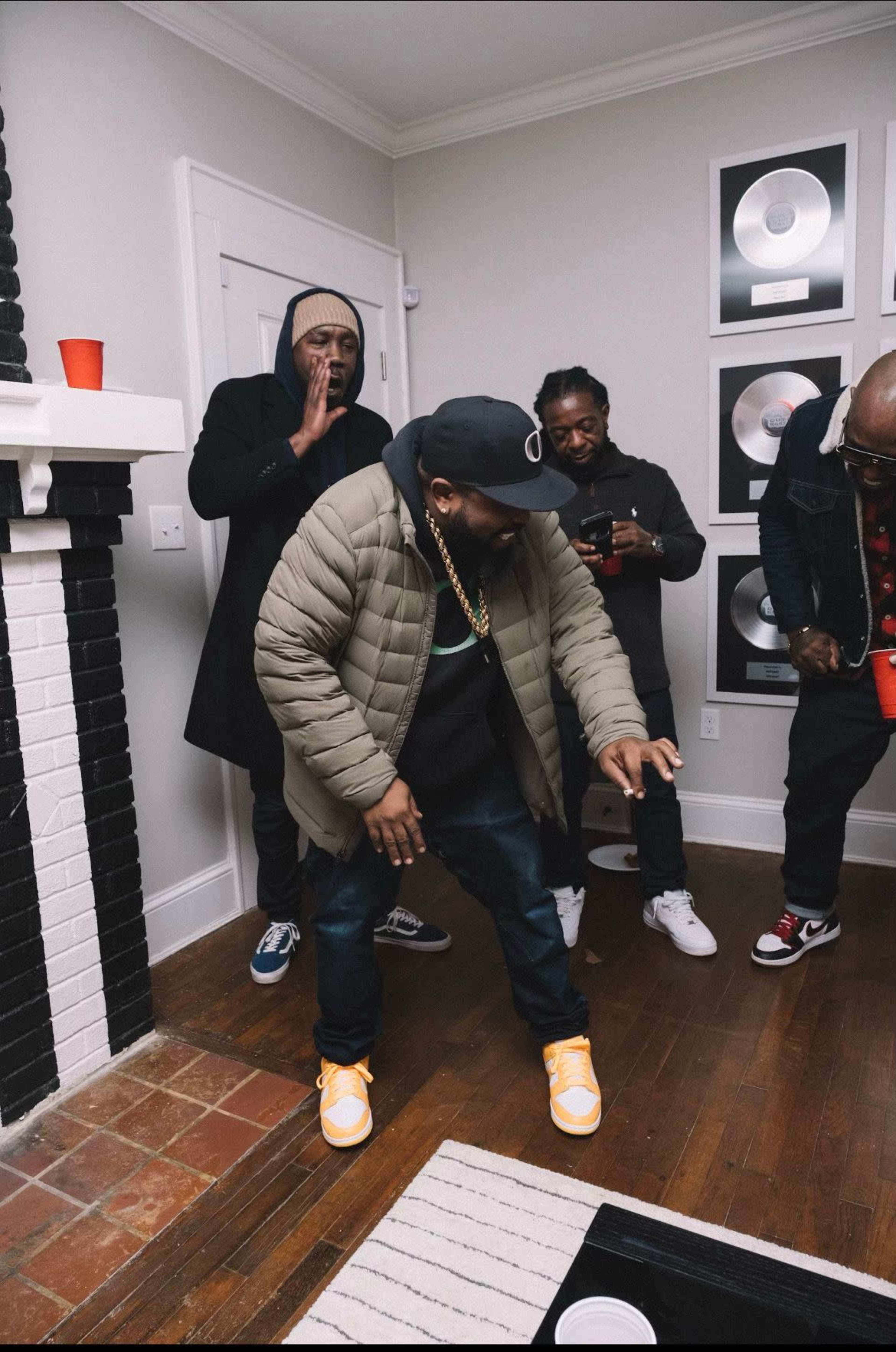 Four men are gathered in a room, with one man dancing while the others observe, and music awards are displayed on the wall.