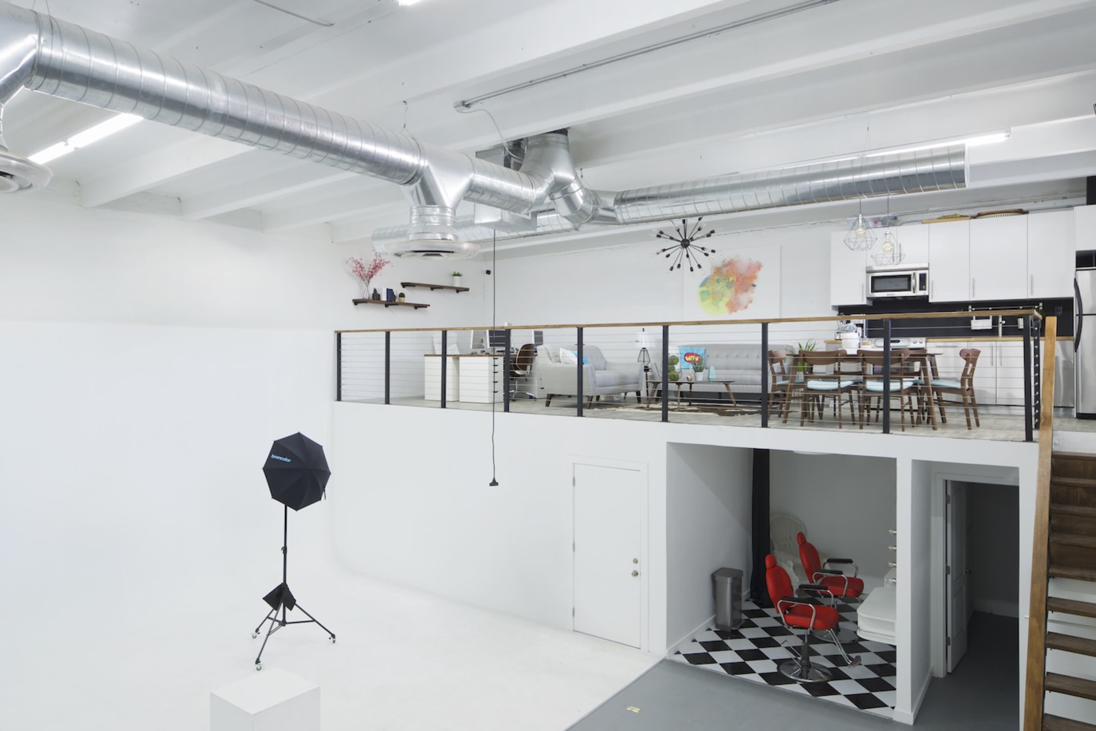 The image shows a spacious, modern studio with a loft area, featuring a seating area and dining space, and a lower level with black and white checkered flooring and two red chairs.