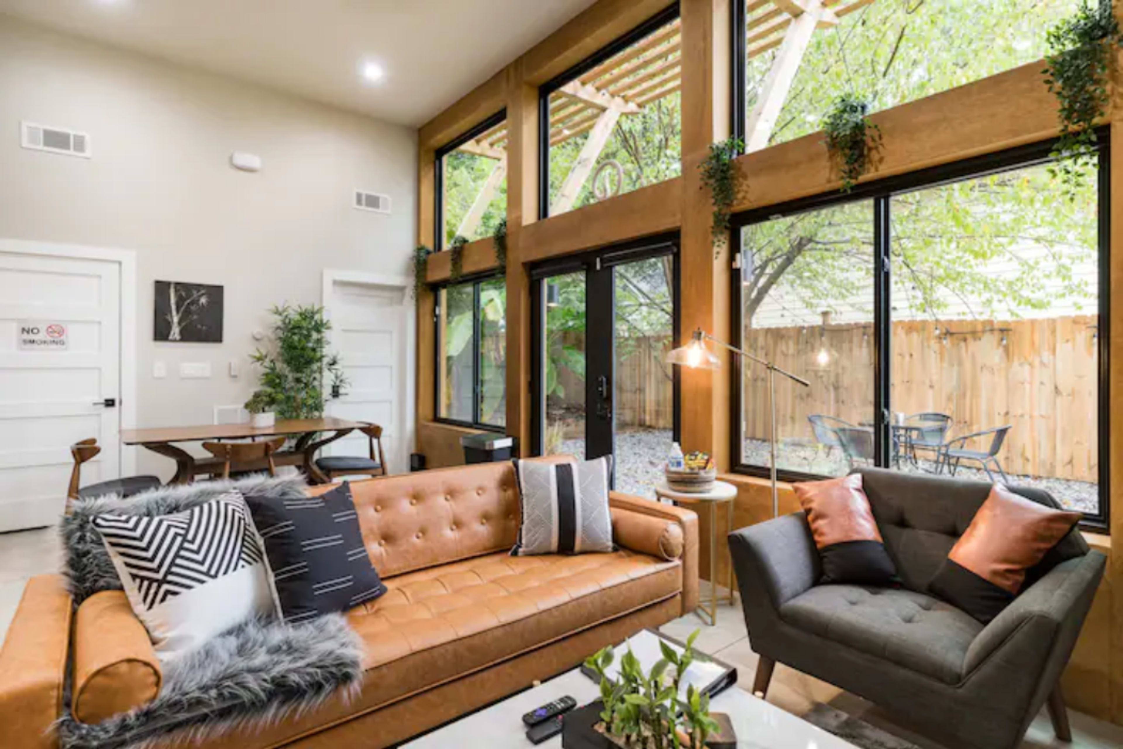 A modern living room with large windows, a brown leather sofa, an angled gray chair, and a view of a fenced outdoor patio with greenery.