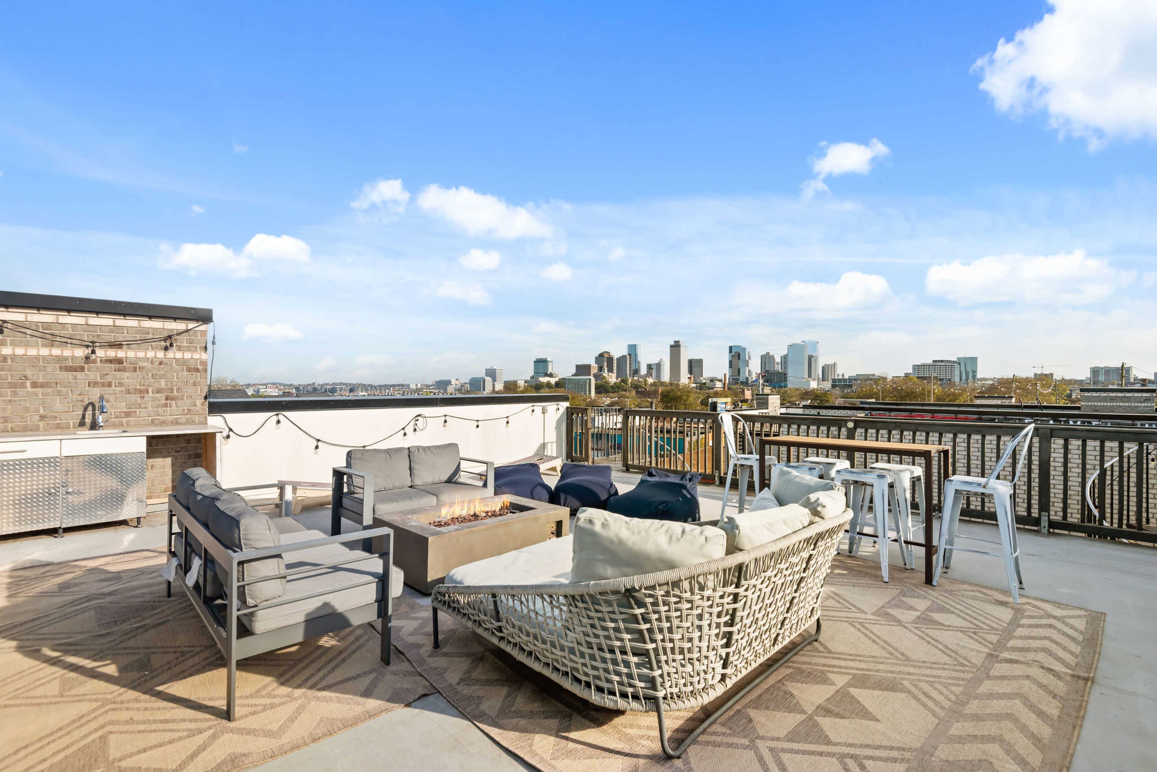 A rooftop terrace features a seating area with a fire pit, surrounded by a city skyline view.
