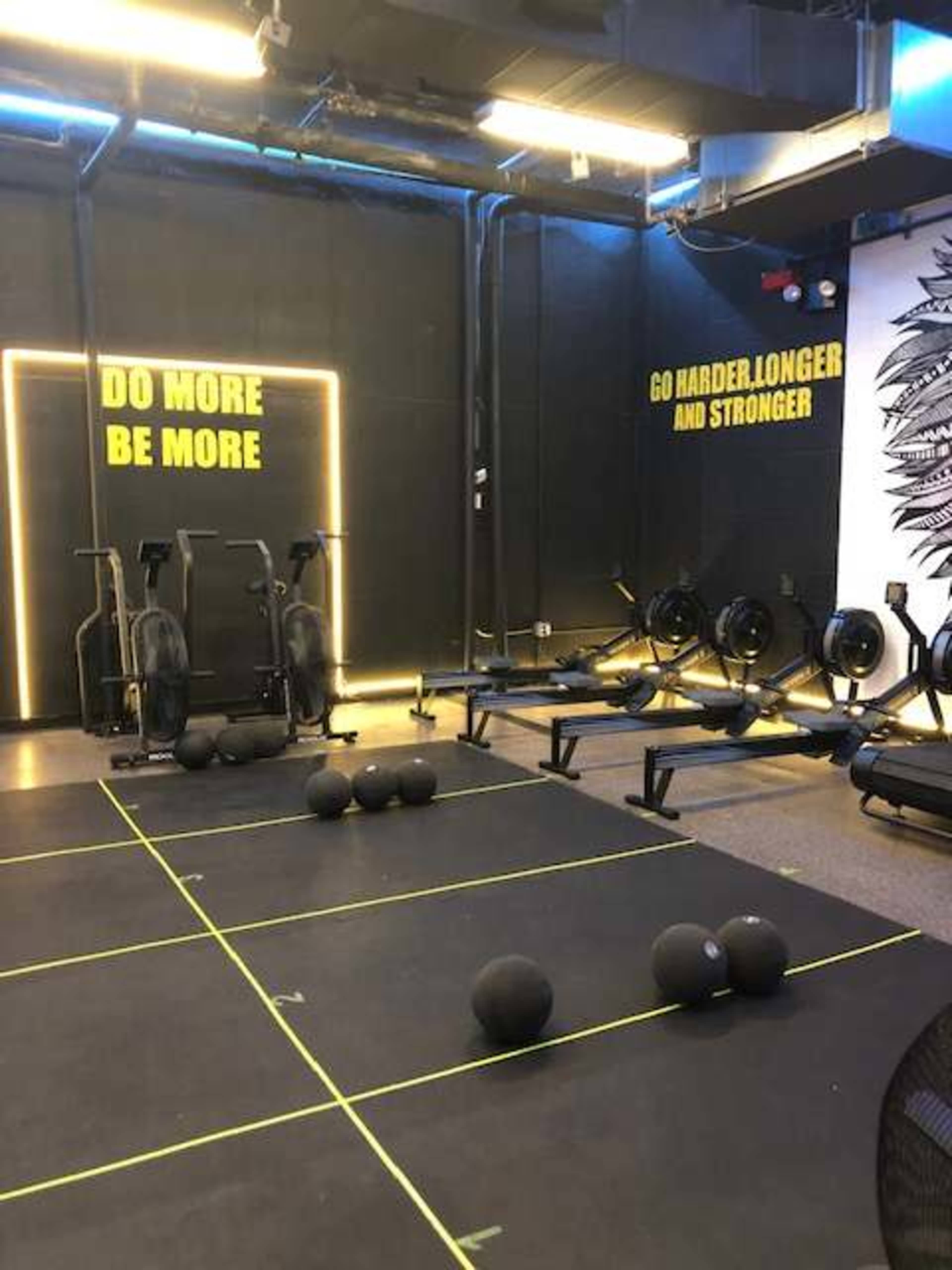 The gym area features exercise equipment including rowers and stationary bikes, with a black wall displaying motivational phrases and medicine balls arranged on the floor.