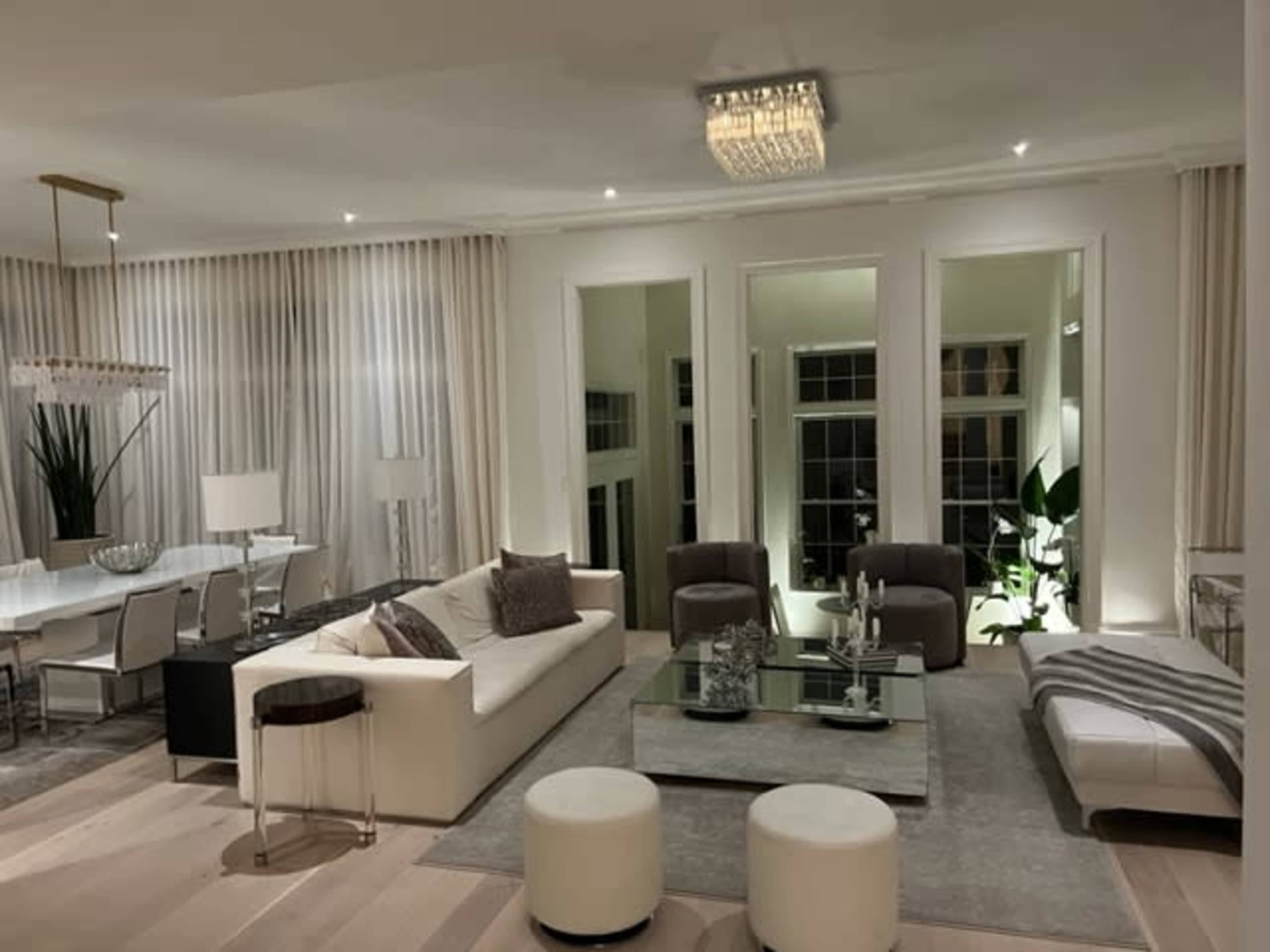 A modern living room with a white sofa, two brown chairs, a glass coffee table, and large windows, complemented by light-colored walls and elegant lighting.