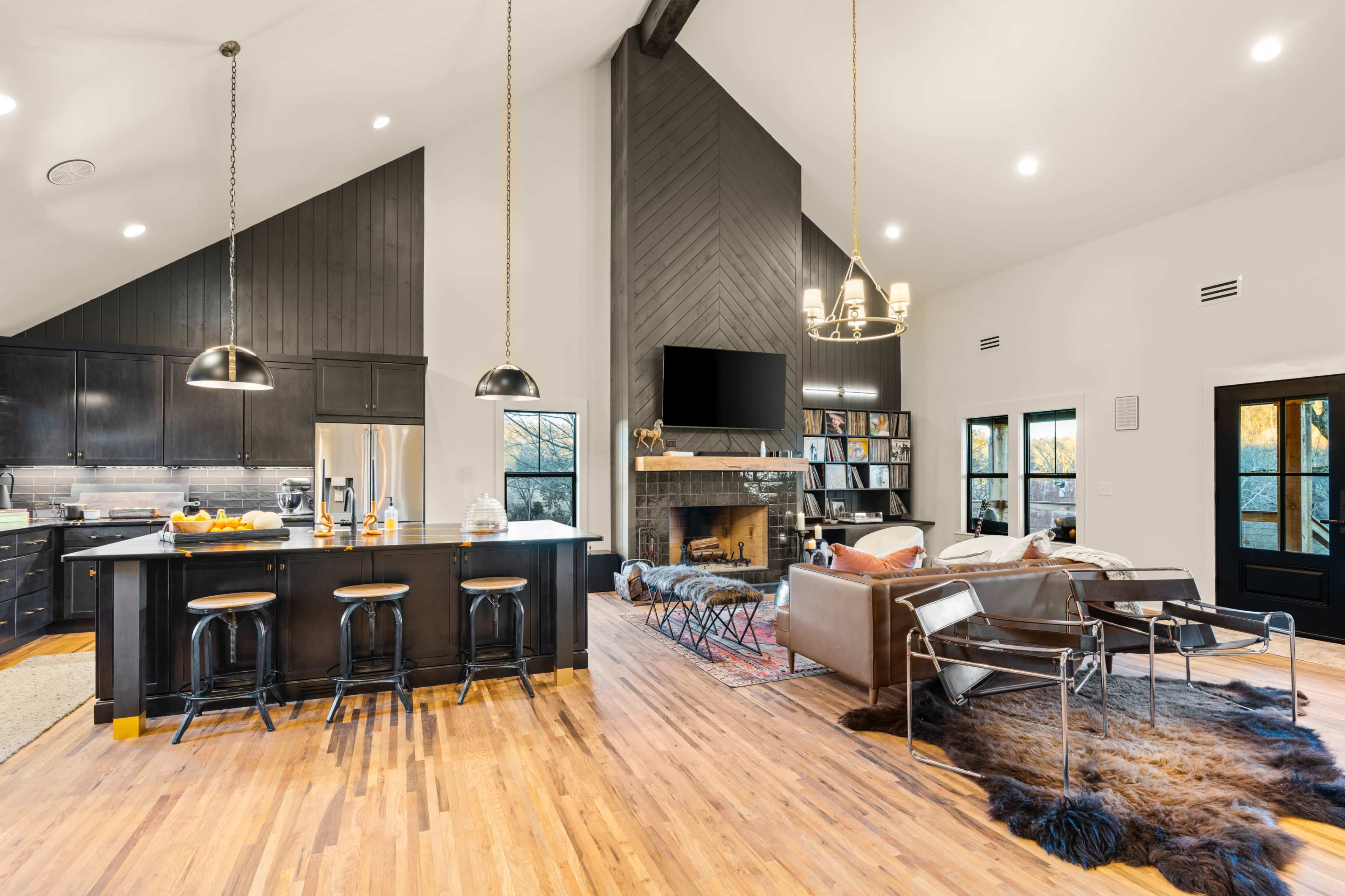 The image shows a modern open-concept living space with a kitchen, dining area, and living room, featuring high ceilings and a fireplace.