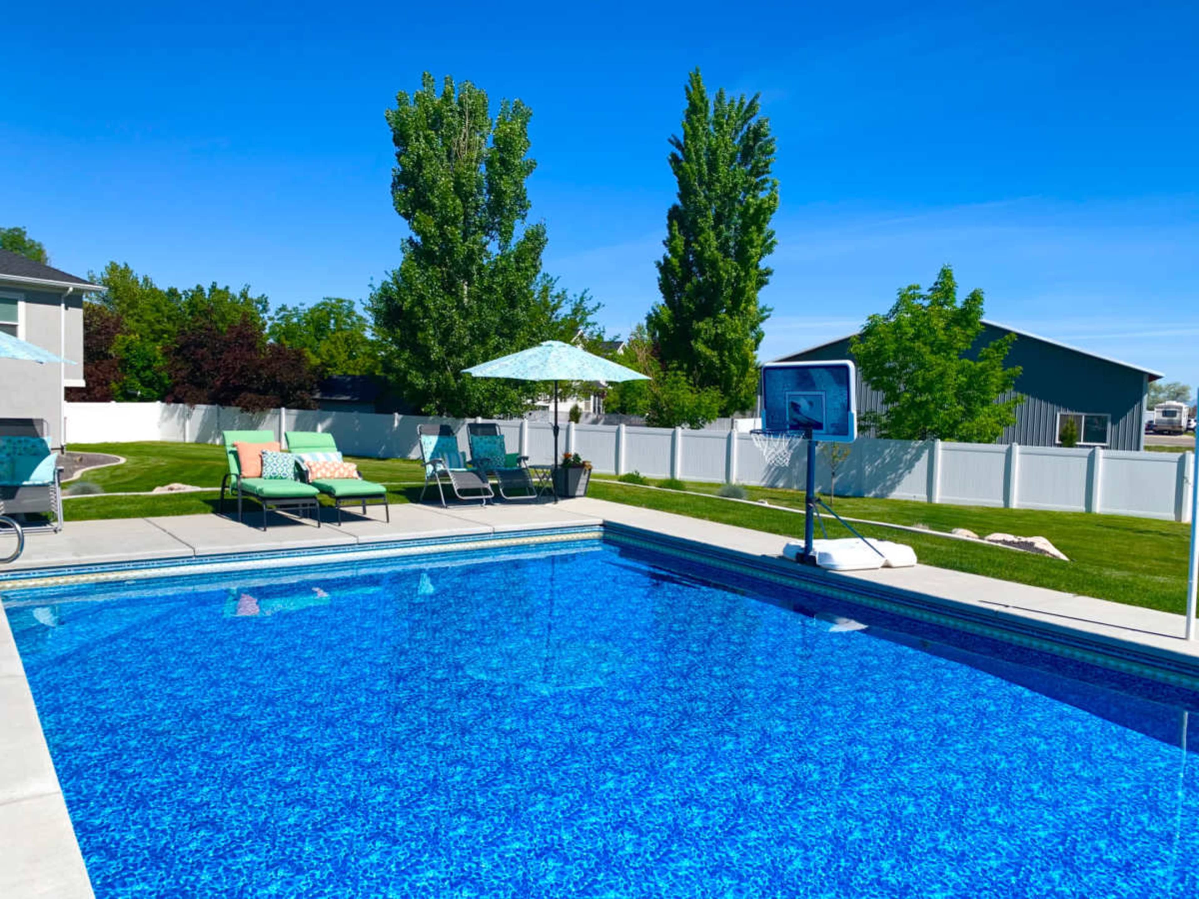 The image shows a clear blue swimming pool surrounded by green lounge chairs and an umbrella, with a basketball hoop positioned nearby and a white fence in the background.