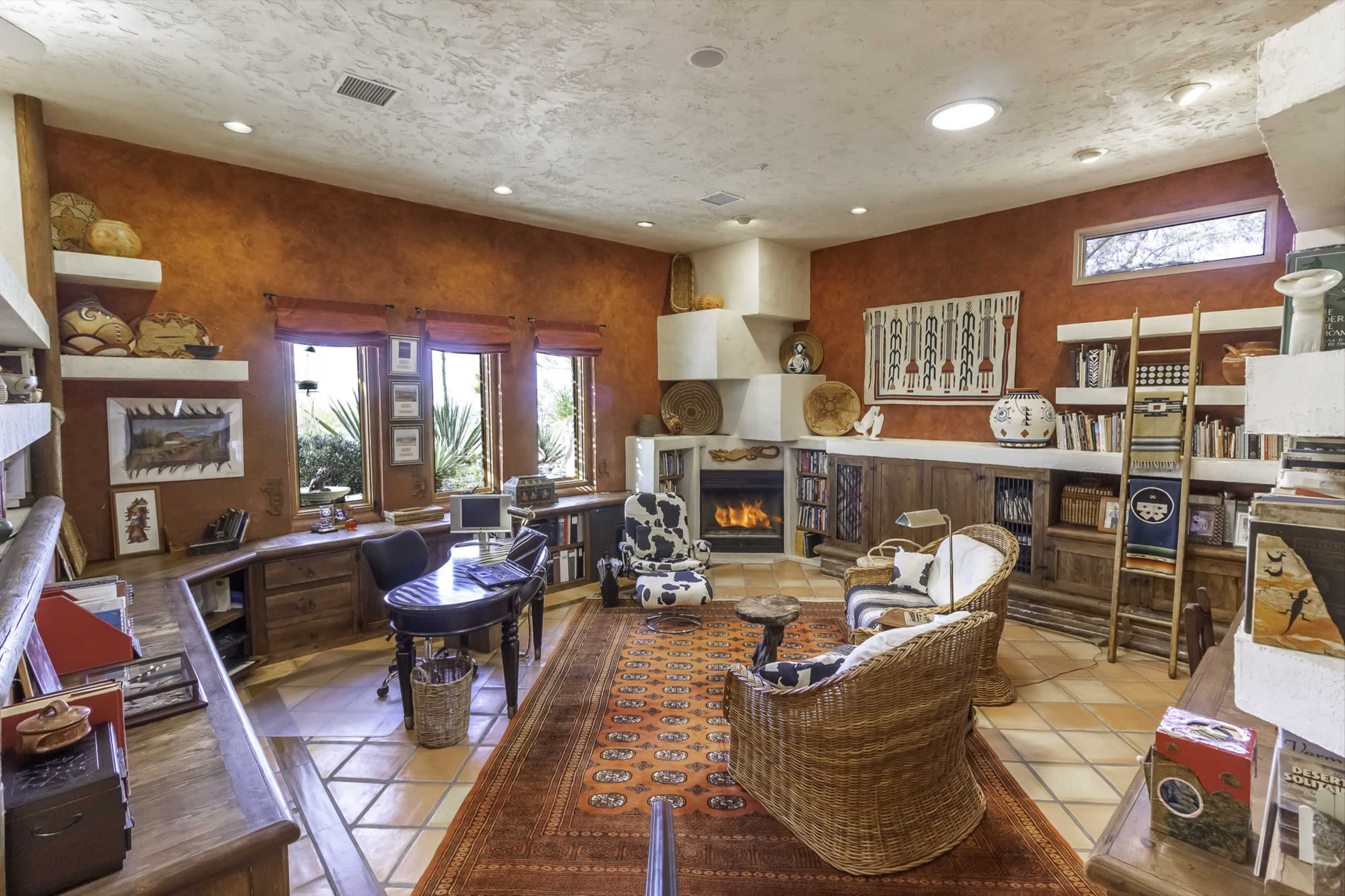 A cozy home office with a round table, a patterned chair, a wicker chair, bookshelves, decorative items, and a fireplace, all arranged around a central area rug.
