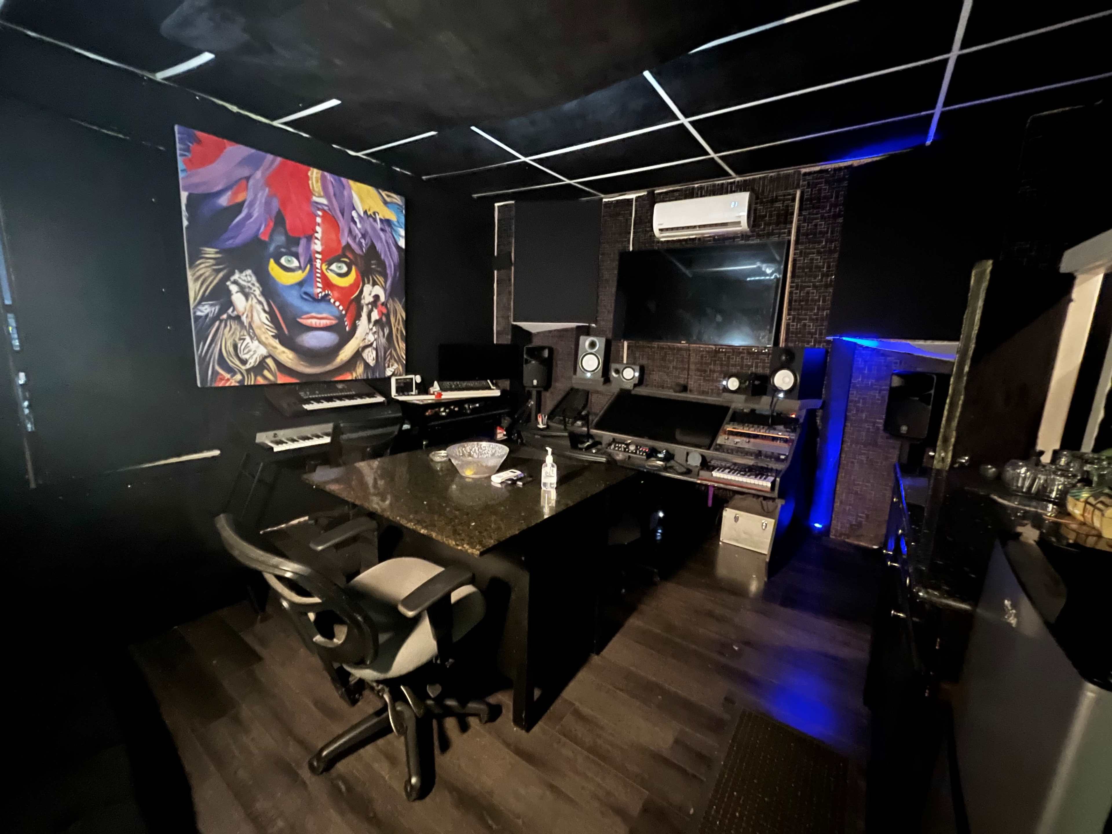 A music studio featuring a central control desk, various electronic instruments, and a large colorful mural on the wall.
