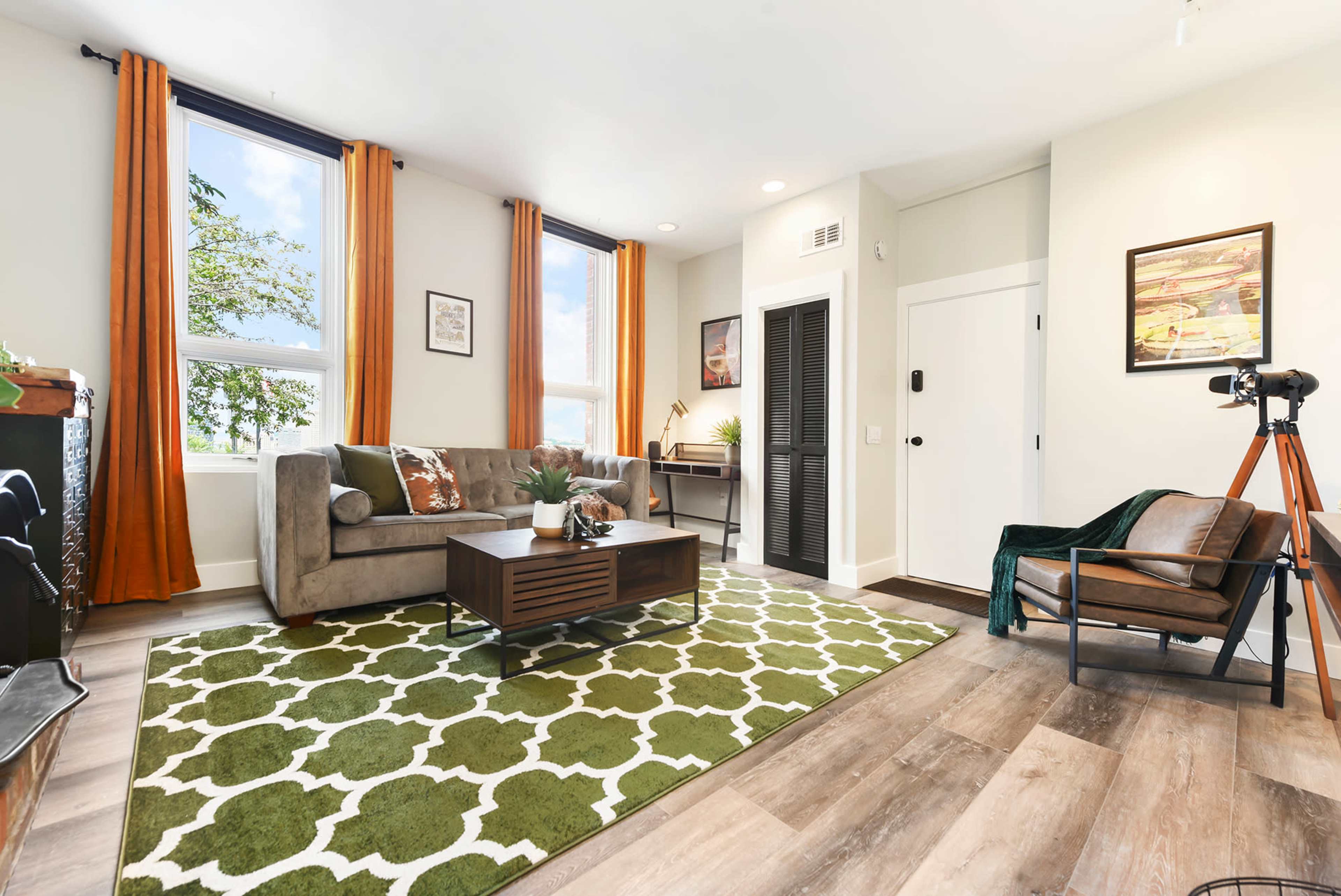 A cozy living room features a gray sofa, a patterned green rug, a coffee table, and a chair beside a window with orange curtains.