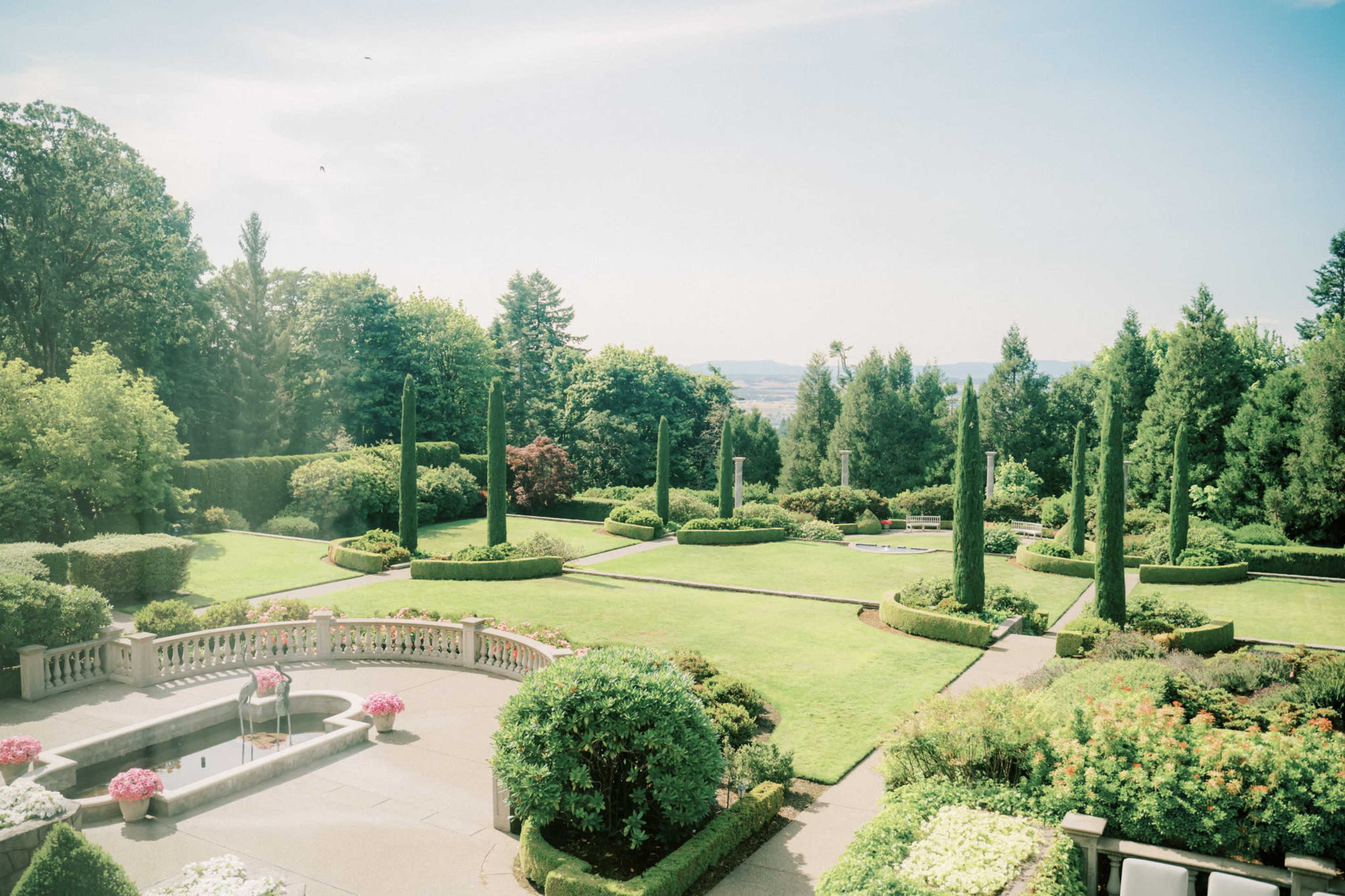 A well-maintained garden featuring symmetrical hedges, tall coniferous trees, and a central fountain surrounded by floral arrangements.