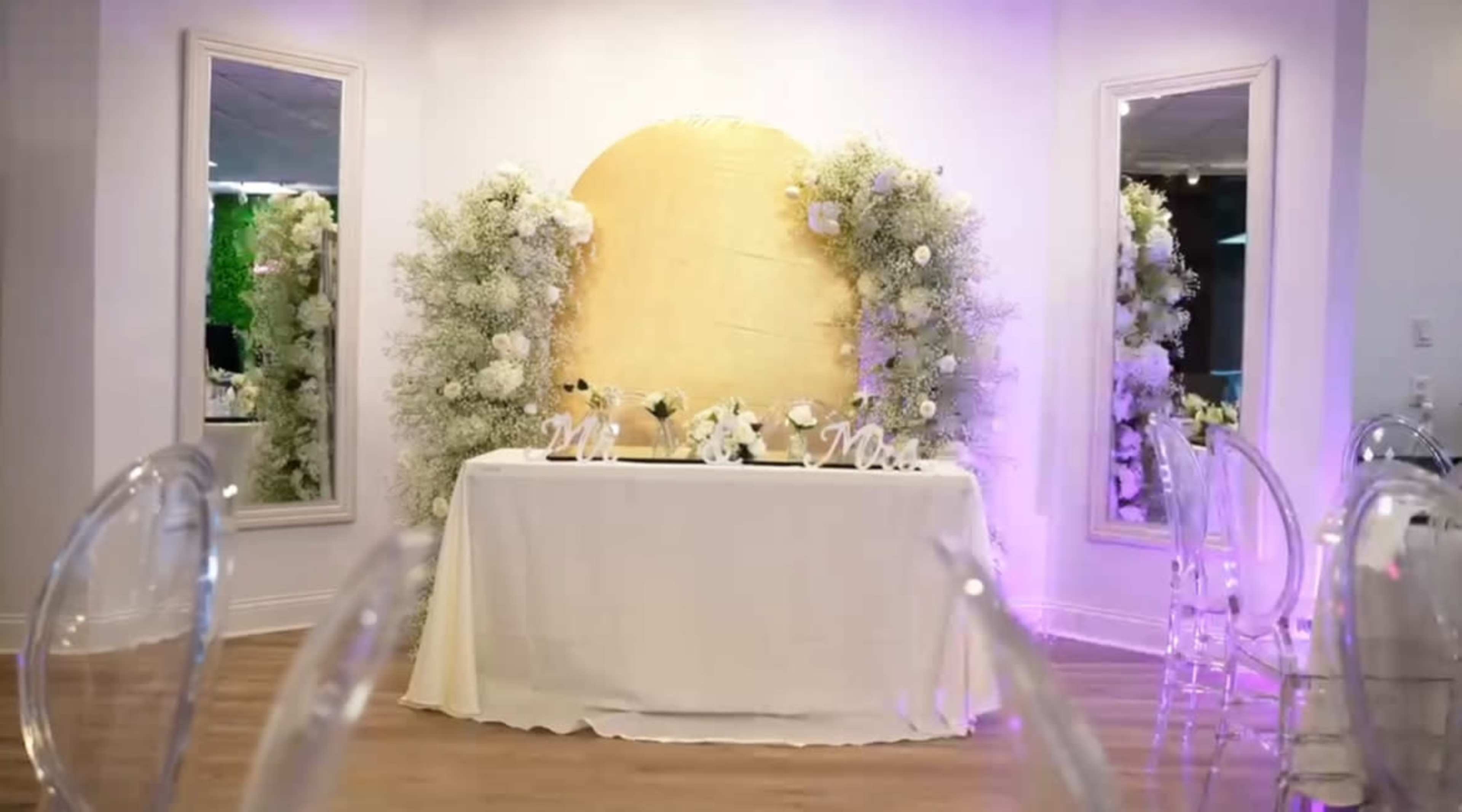 A decorated event space with a golden backdrop, floral arrangements, and a table set with white cloth and decorative items.