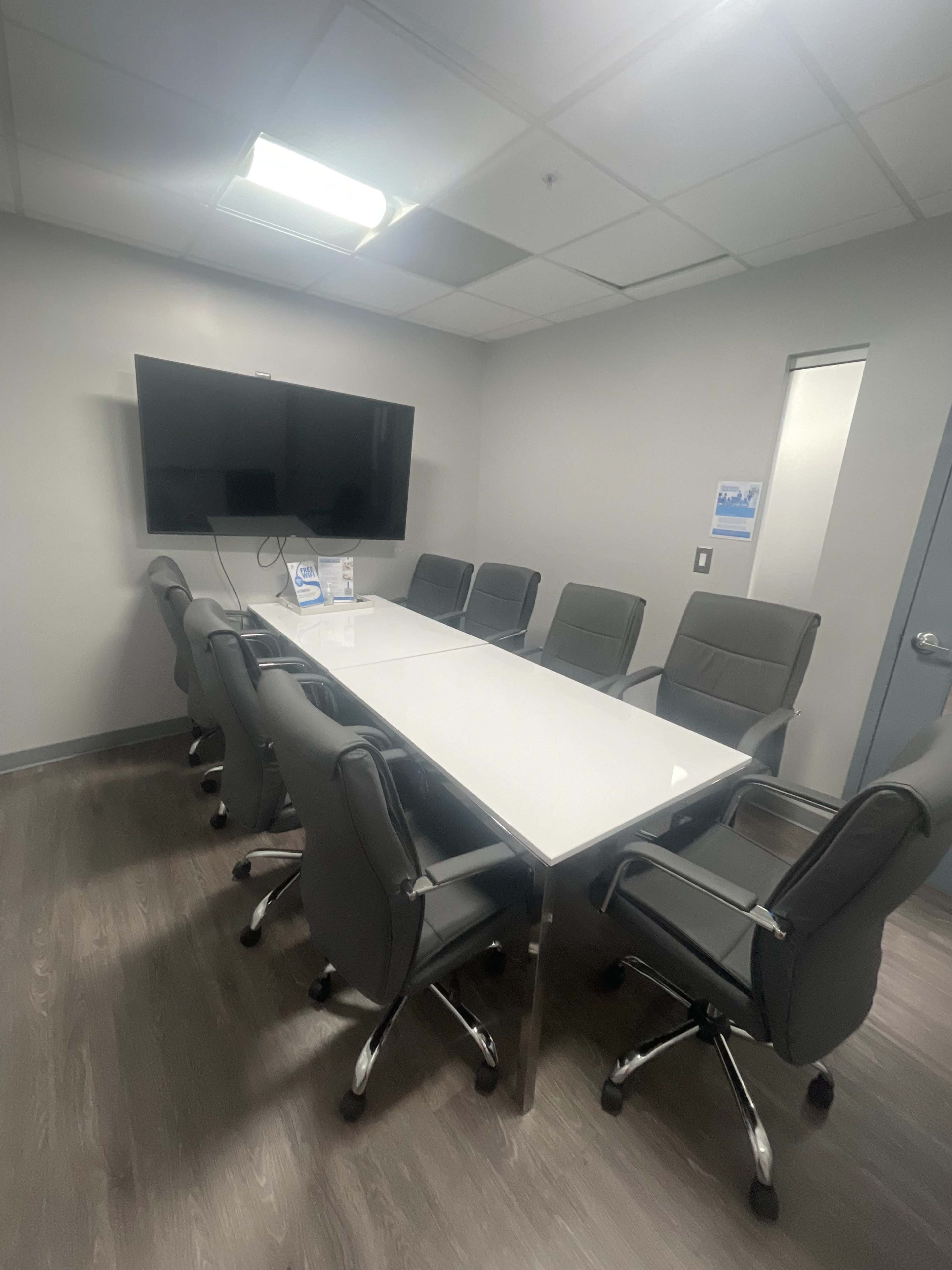 A modern conference room features a large white table surrounded by eight gray chairs and a wall-mounted television.