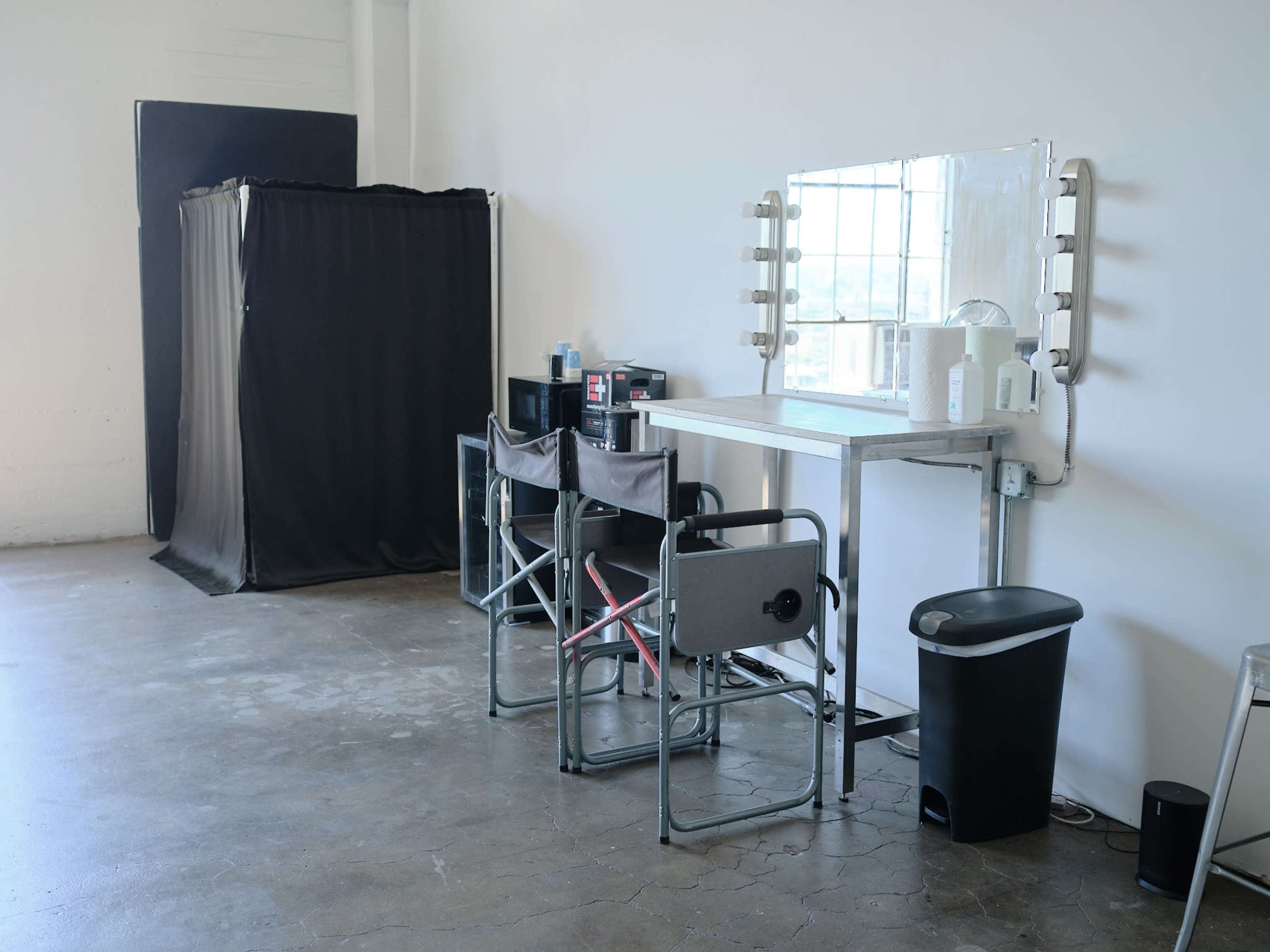 A minimalist makeup station with a mirror and lights, accompanied by several folding chairs and a privacy curtain in an industrial-style room.