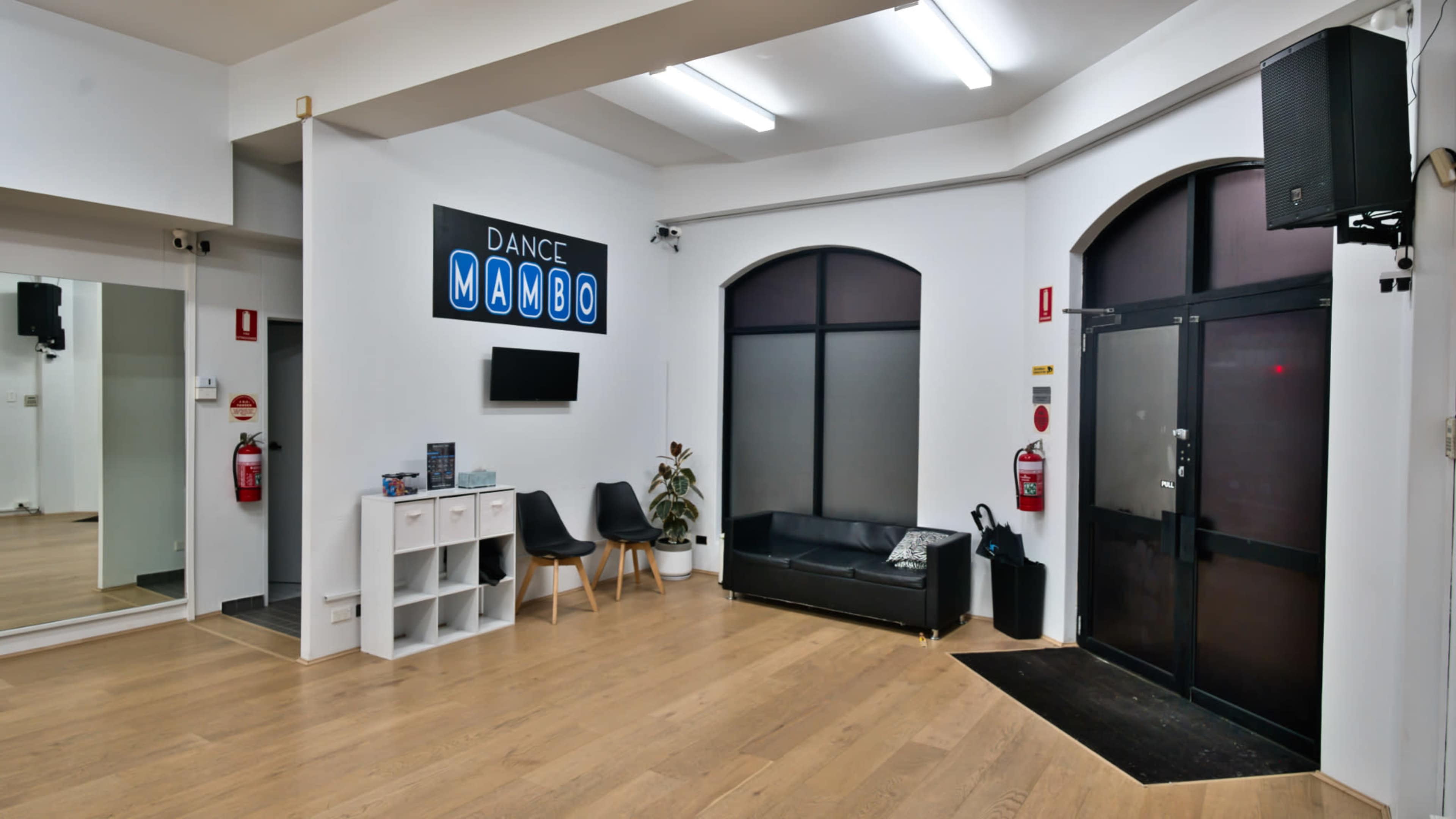 The image shows a dance studio entrance with a reception area featuring a black couch, a small table, and a mirrored wall, under bright overhead lighting and a sign that reads "DANCE MAMBO."