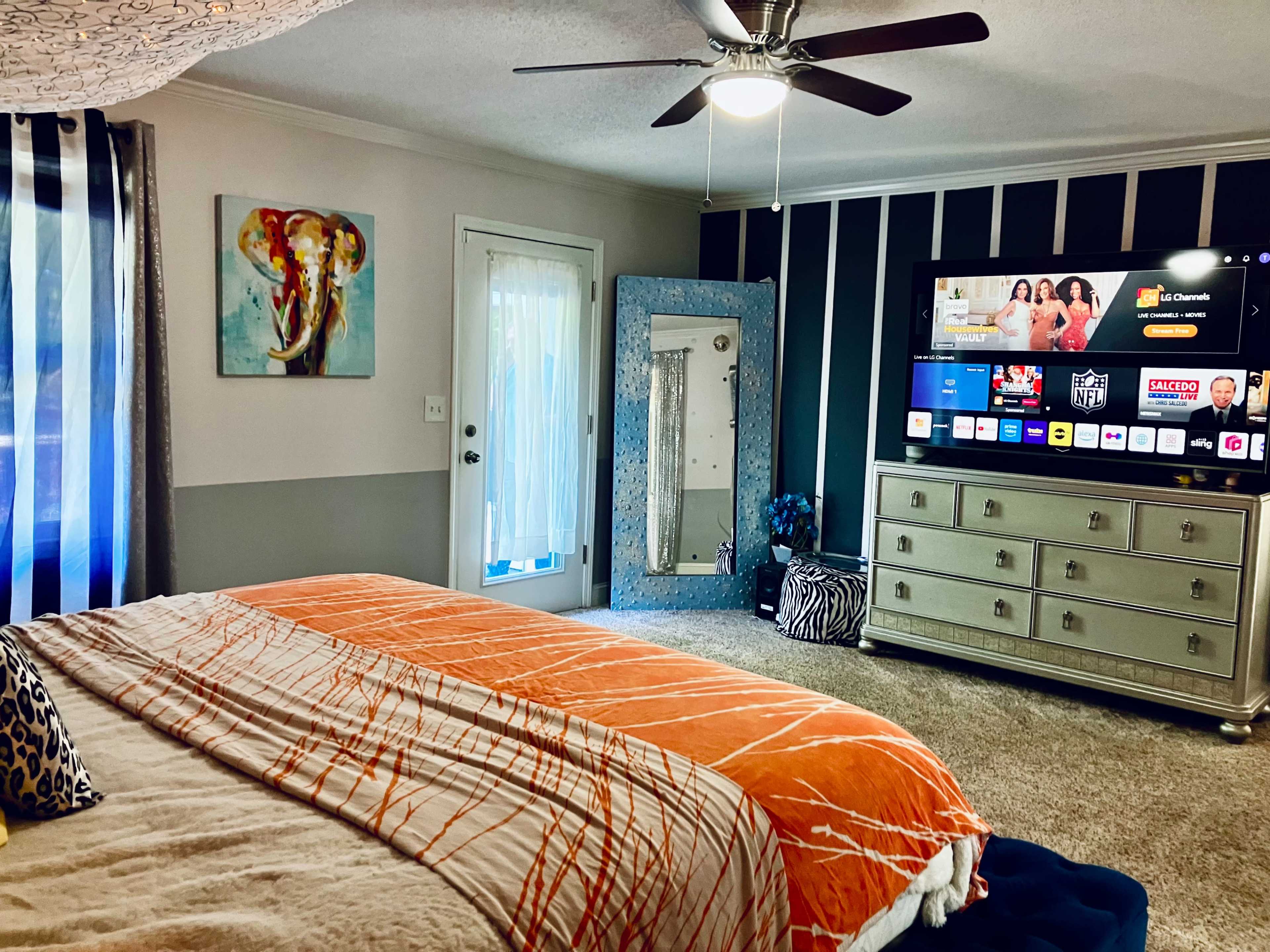 The image shows a bedroom featuring a bed with an orange and cream-colored blanket, a TV mounted on the wall with various streaming options displayed, and an elephant painting hanging beside a mirror and a door leading outside.