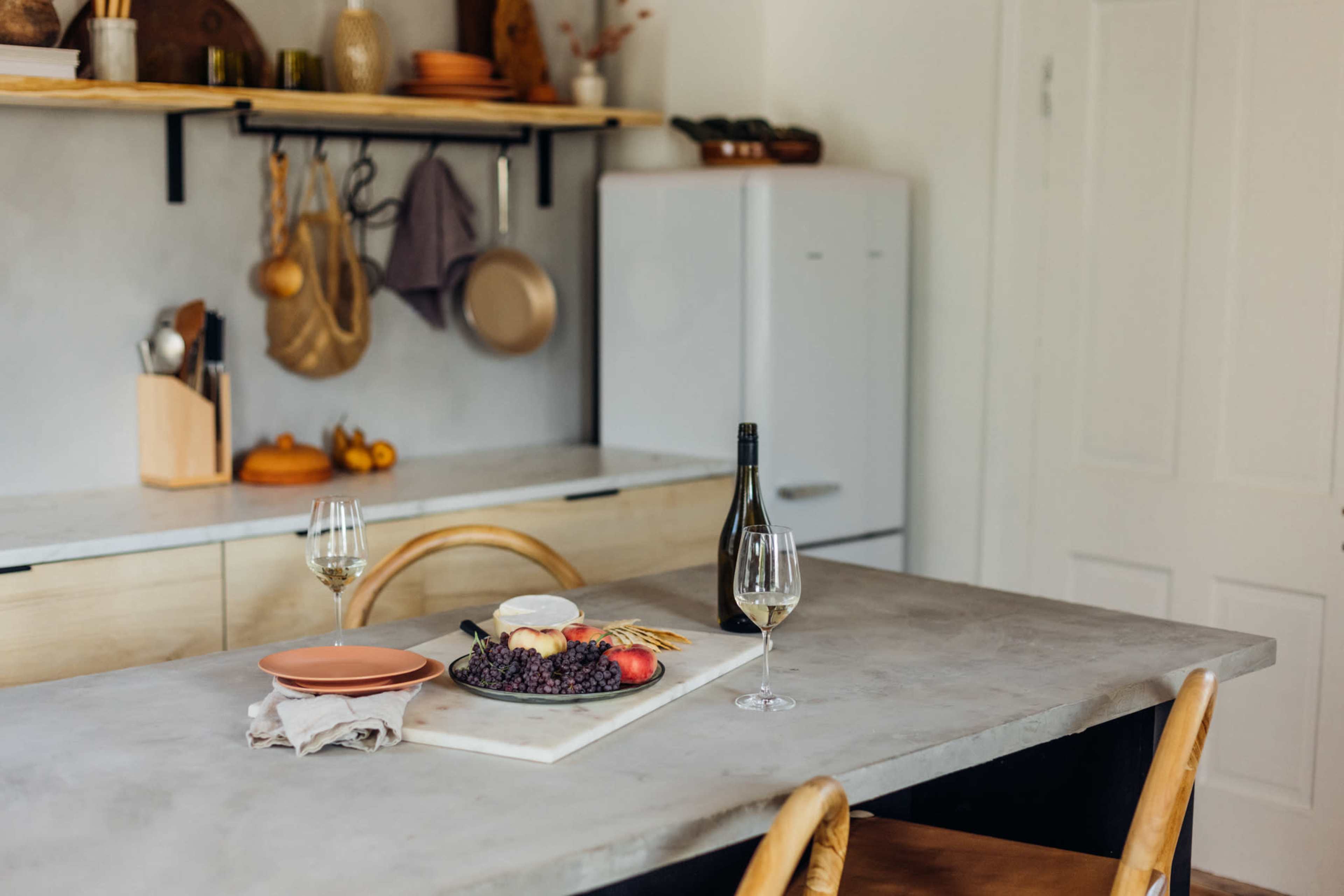A dining table is set with a platter of grapes, an apple, and a bottle of wine, surrounded by minimalist kitchen decor and shelves.