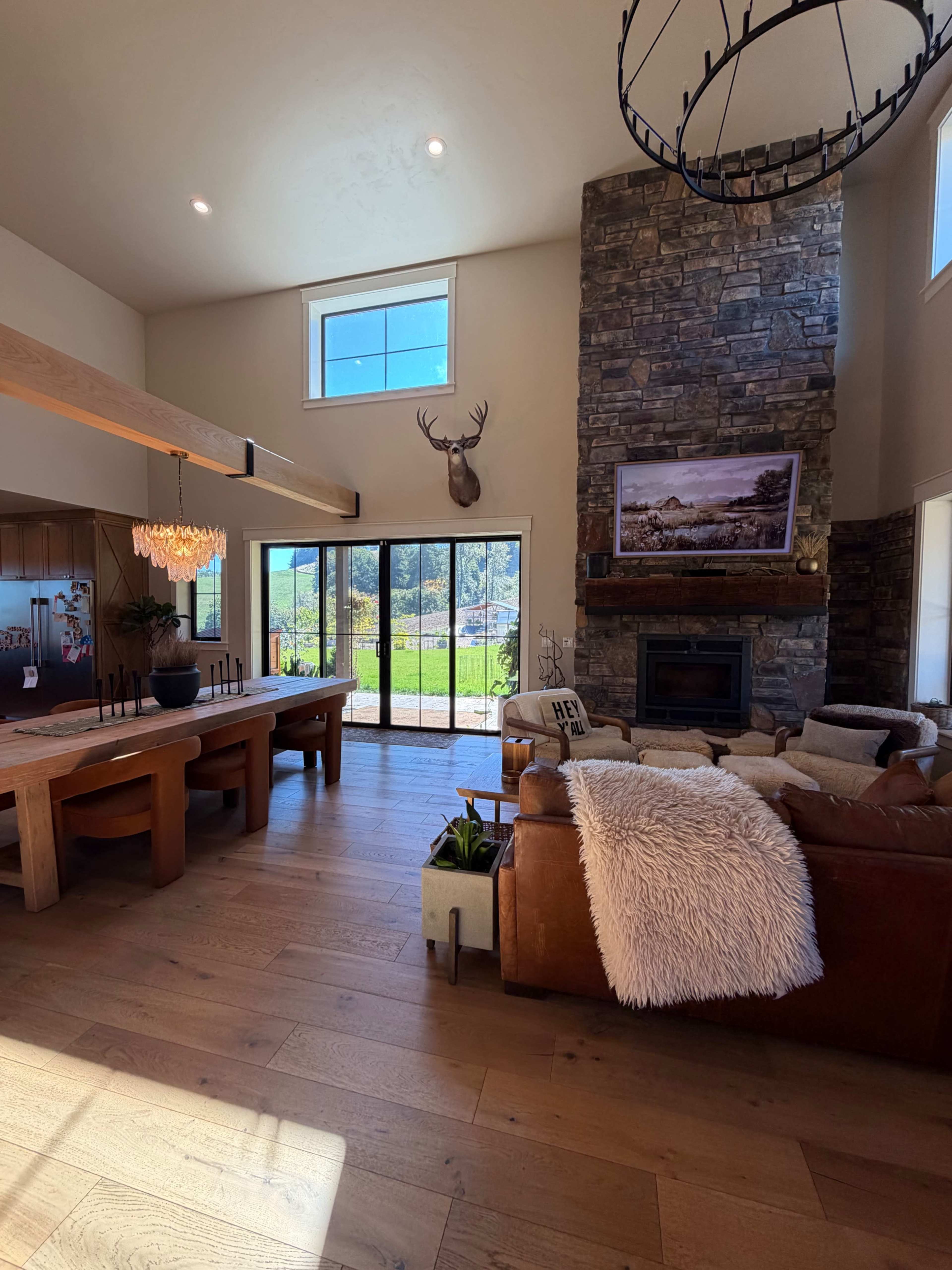The image shows a spacious living area featuring a stone fireplace, a large window with a view of the outdoors, a wood dining table, and a leather sofa adorned with a throw blanket.