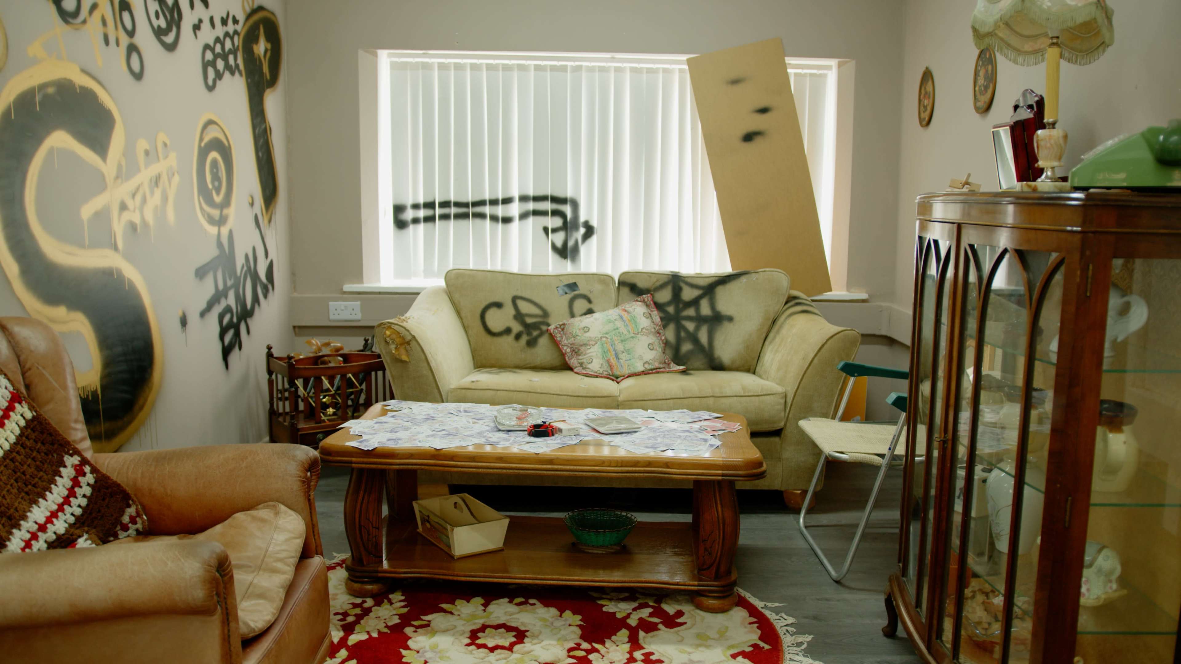 The room features a worn sofa and armchair, a coffee table cluttered with papers, and walls adorned with graffiti, alongside a leaning piece of plywood.
