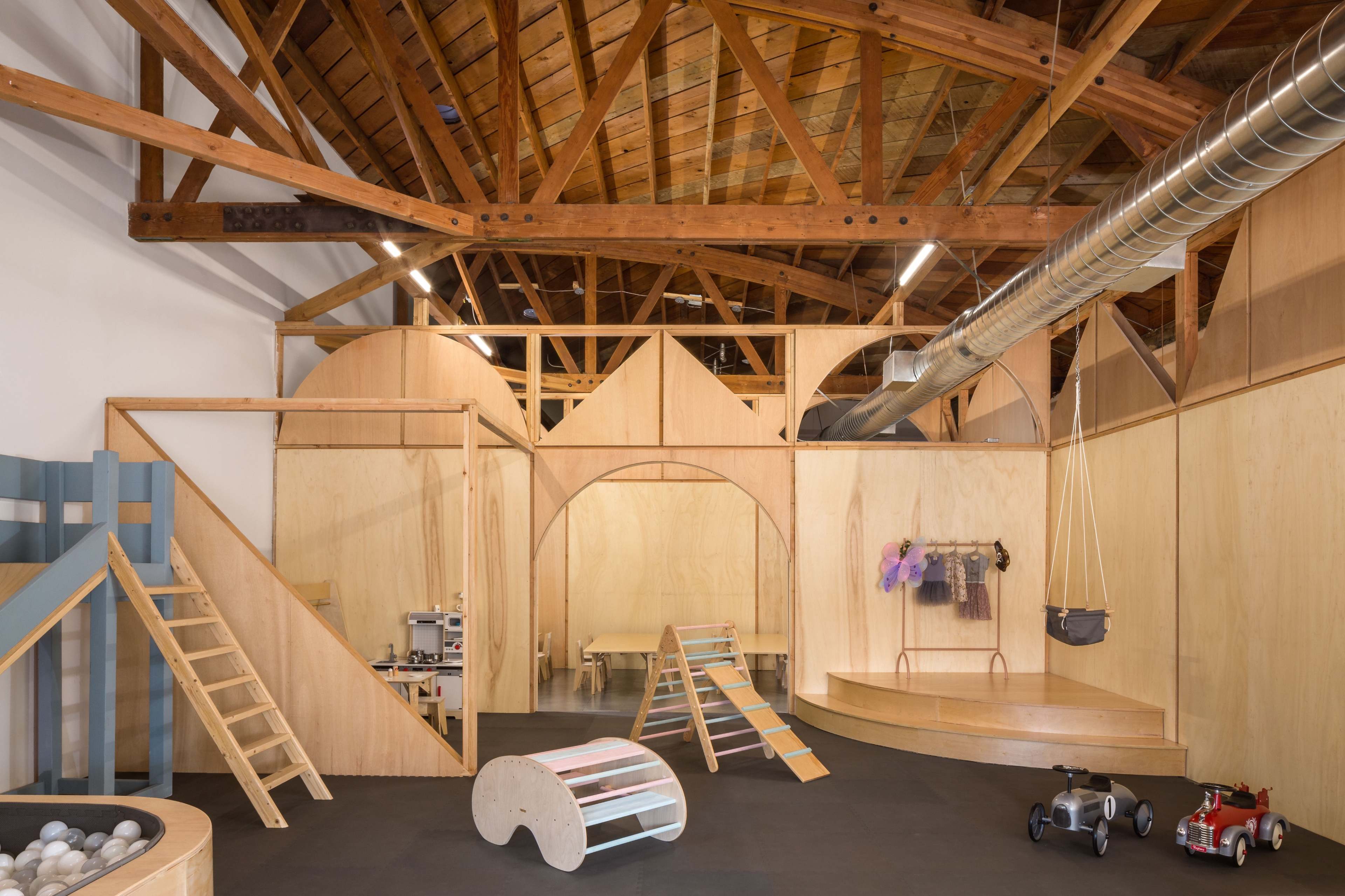 A wooden play area featuring ramps, slides, and various toys, with a high ceiling supported by exposed beams.