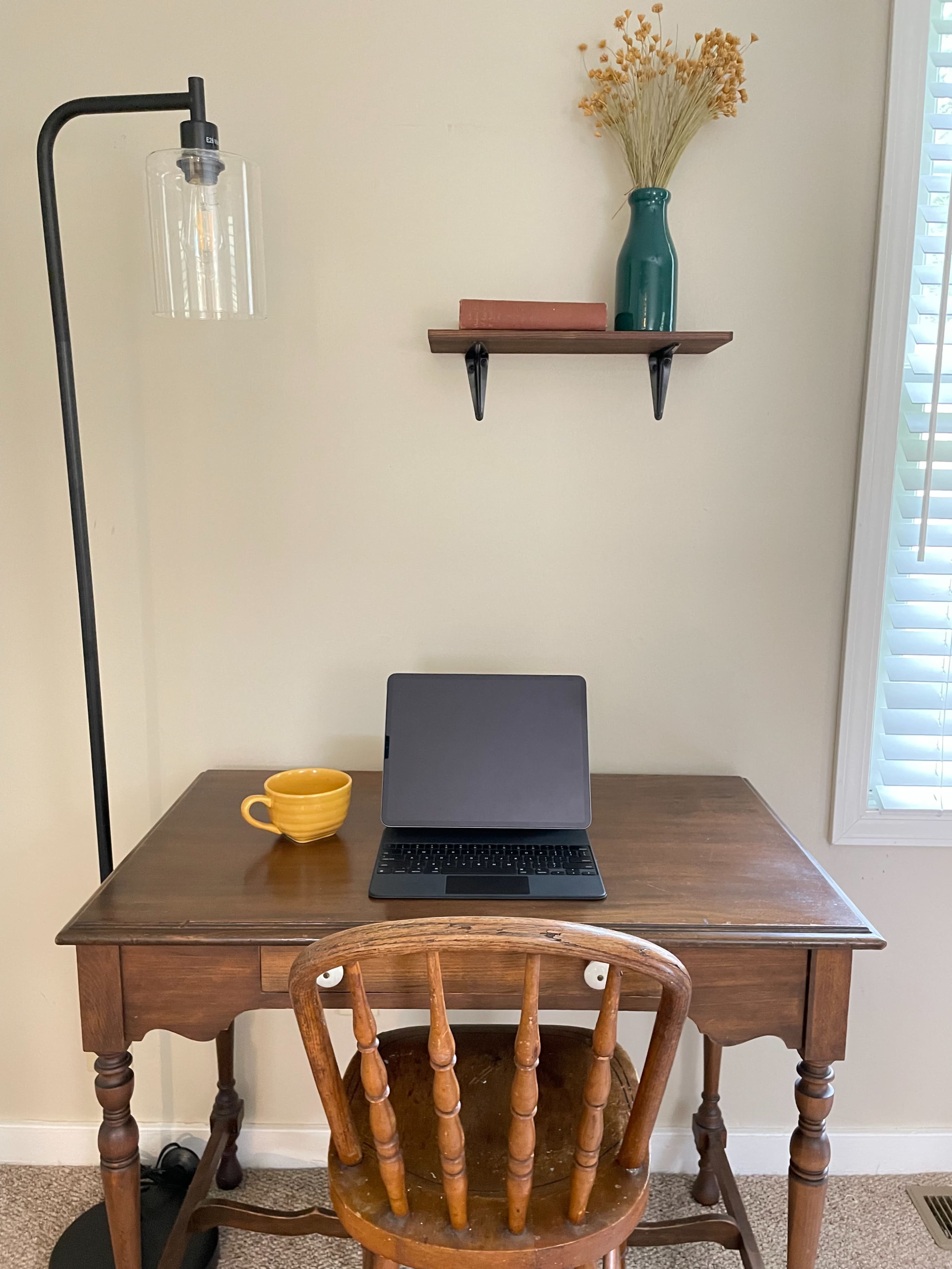 A wooden desk with a chair, a laptop, a yellow cup, and a shelf with a vase and books is positioned against a wall.