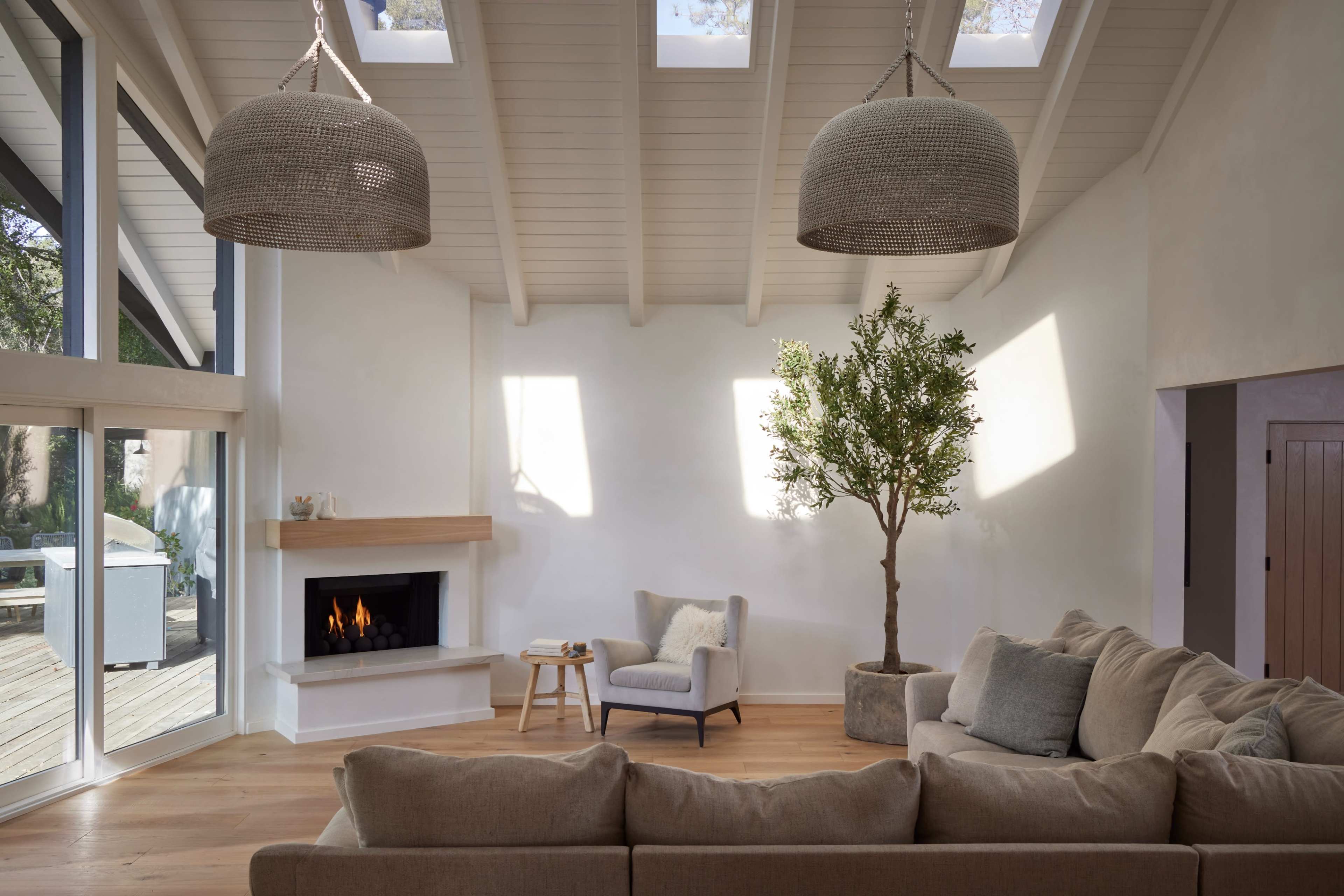 The image shows a modern living room with a large sectional sofa, a small armchair, a fireplace, and a potted tree beneath a vaulted ceiling featuring skylights.