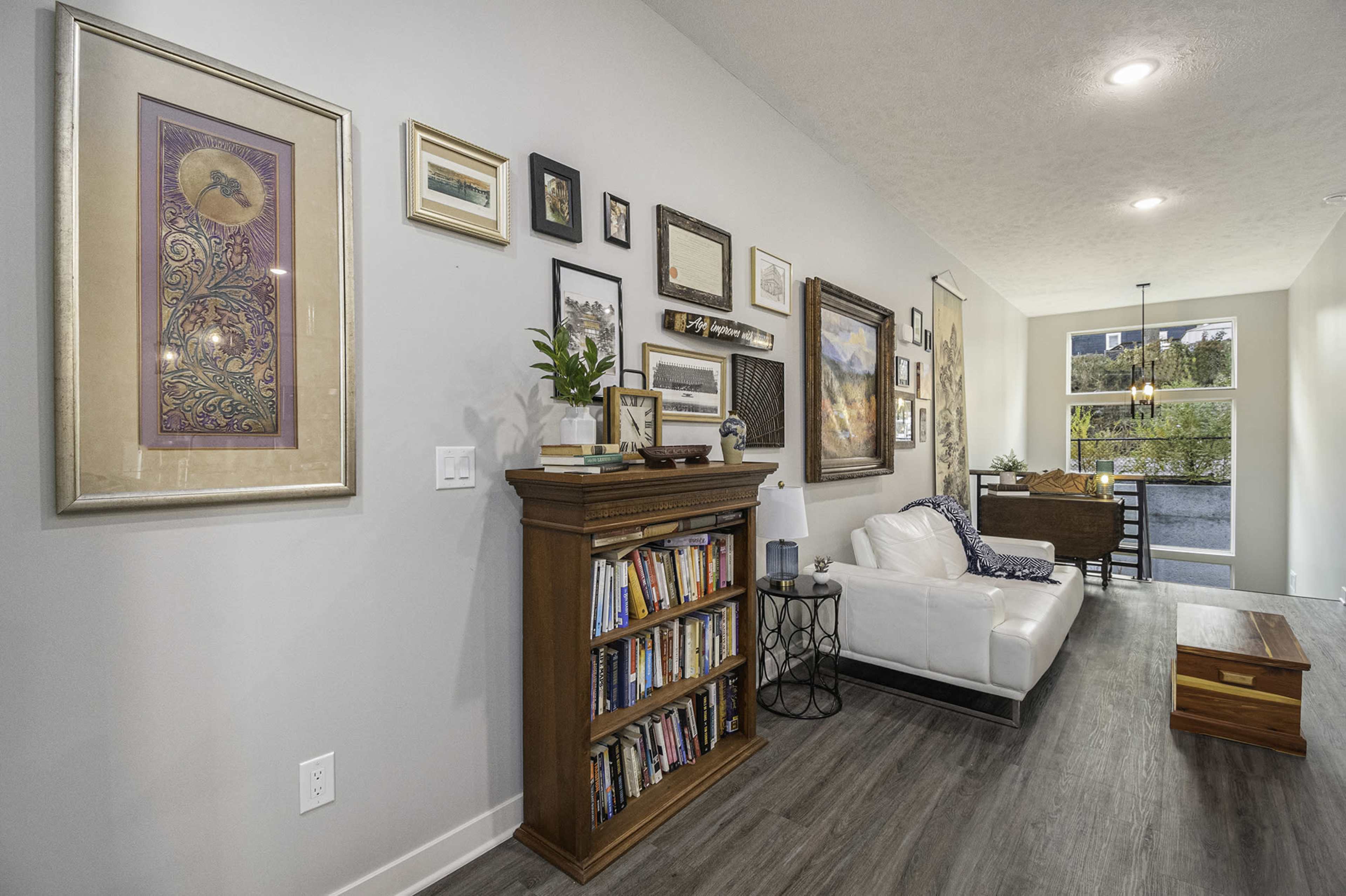 A modern living room with a white sofa, a wooden bookshelf filled with books, and various framed artworks on the walls.