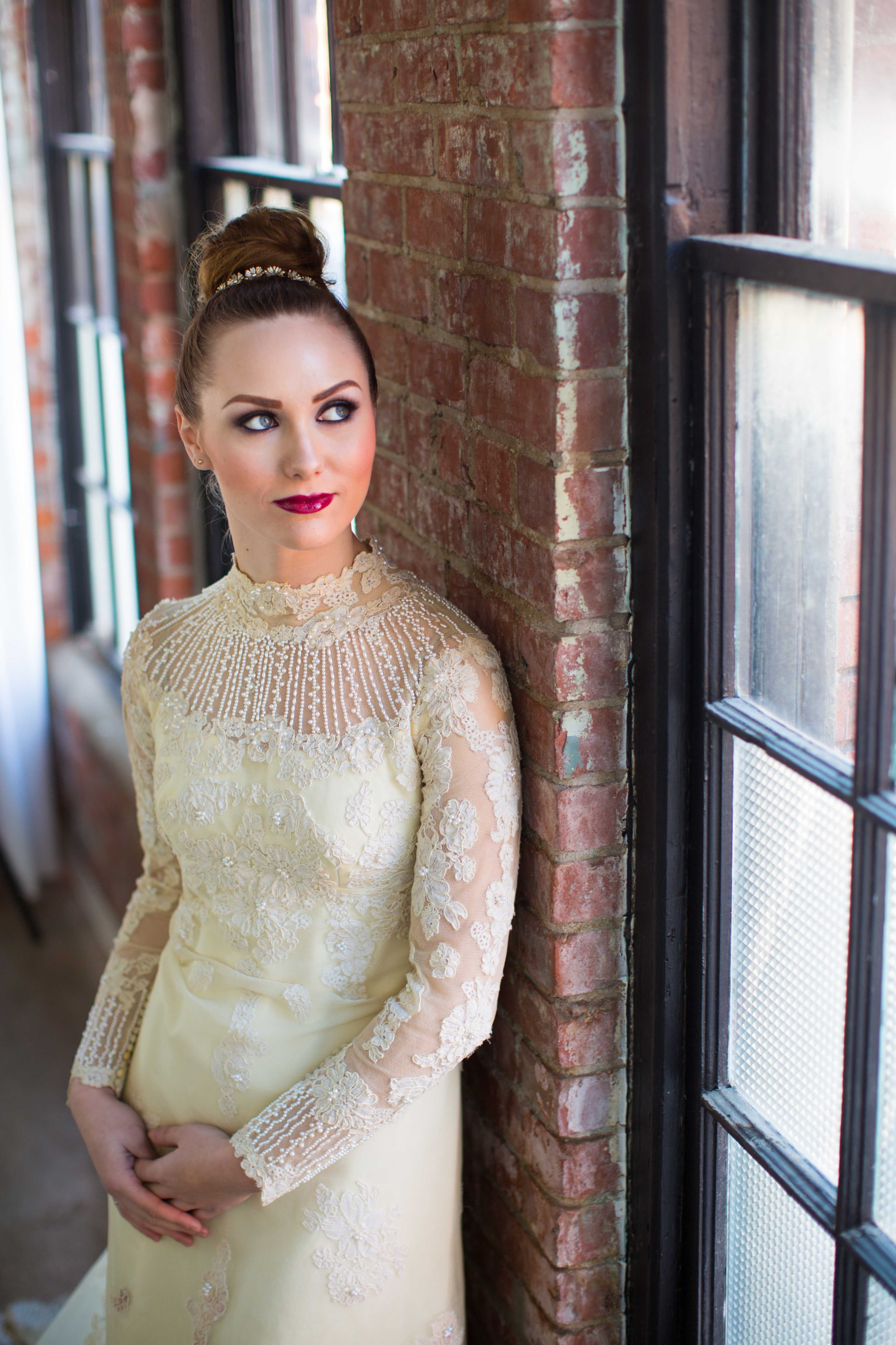 A woman in a yellow gown with lace sleeves poses by a brick wall and large windows.