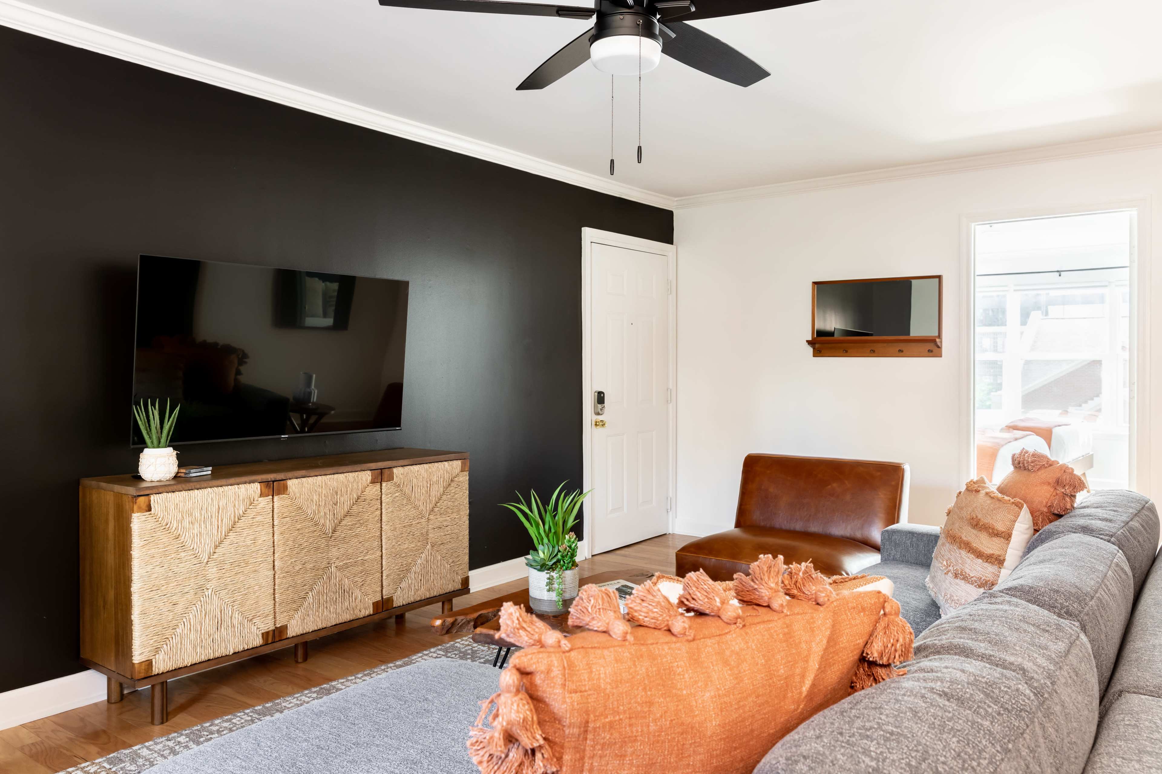 The living room features a black accent wall, a large flat-screen TV on a wooden console, a brown leather chair, and a gray sofa adorned with orange cushions.