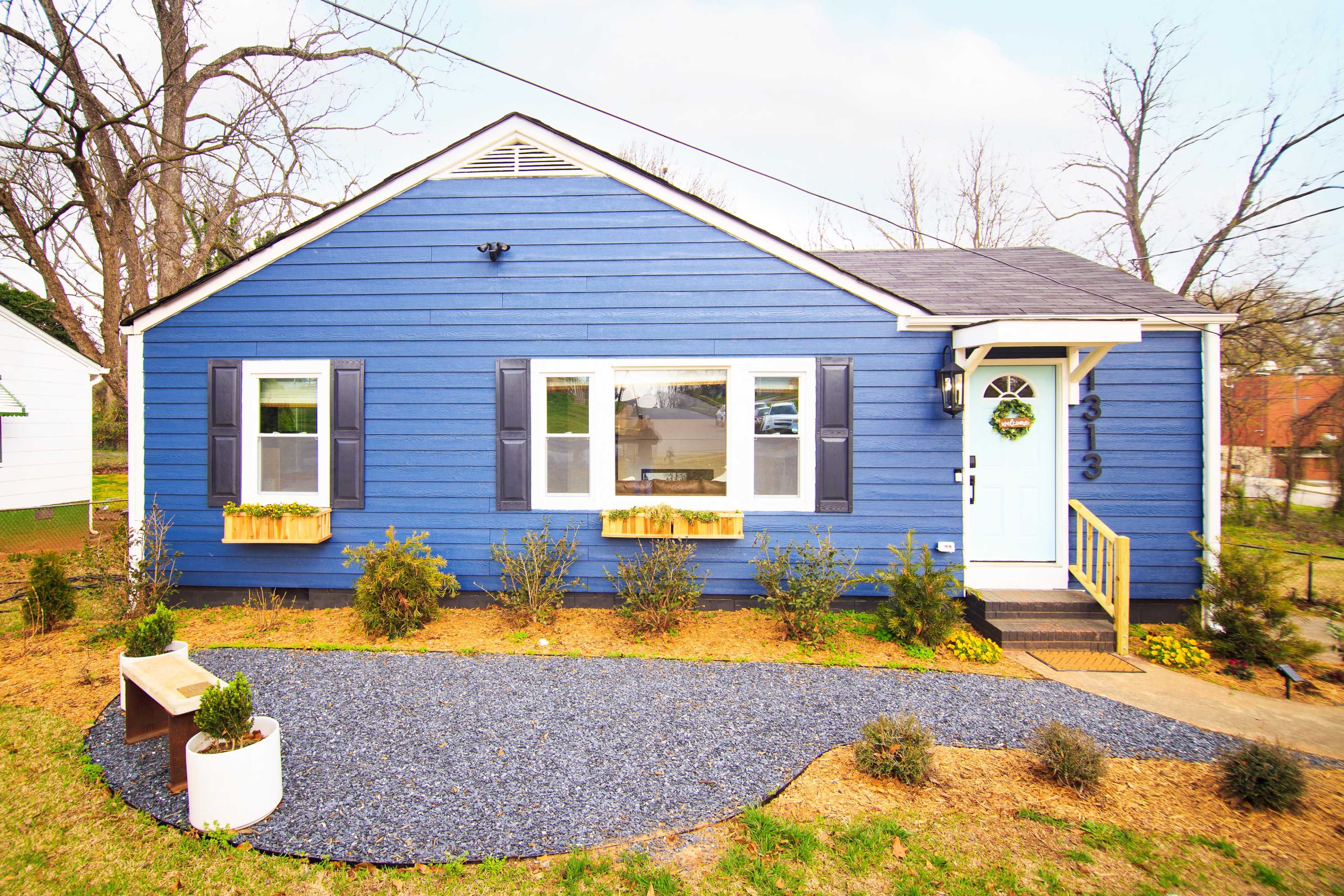 The image shows a blue, single-story house with white trim, a front porch, and small shrubs along a gravel path.
