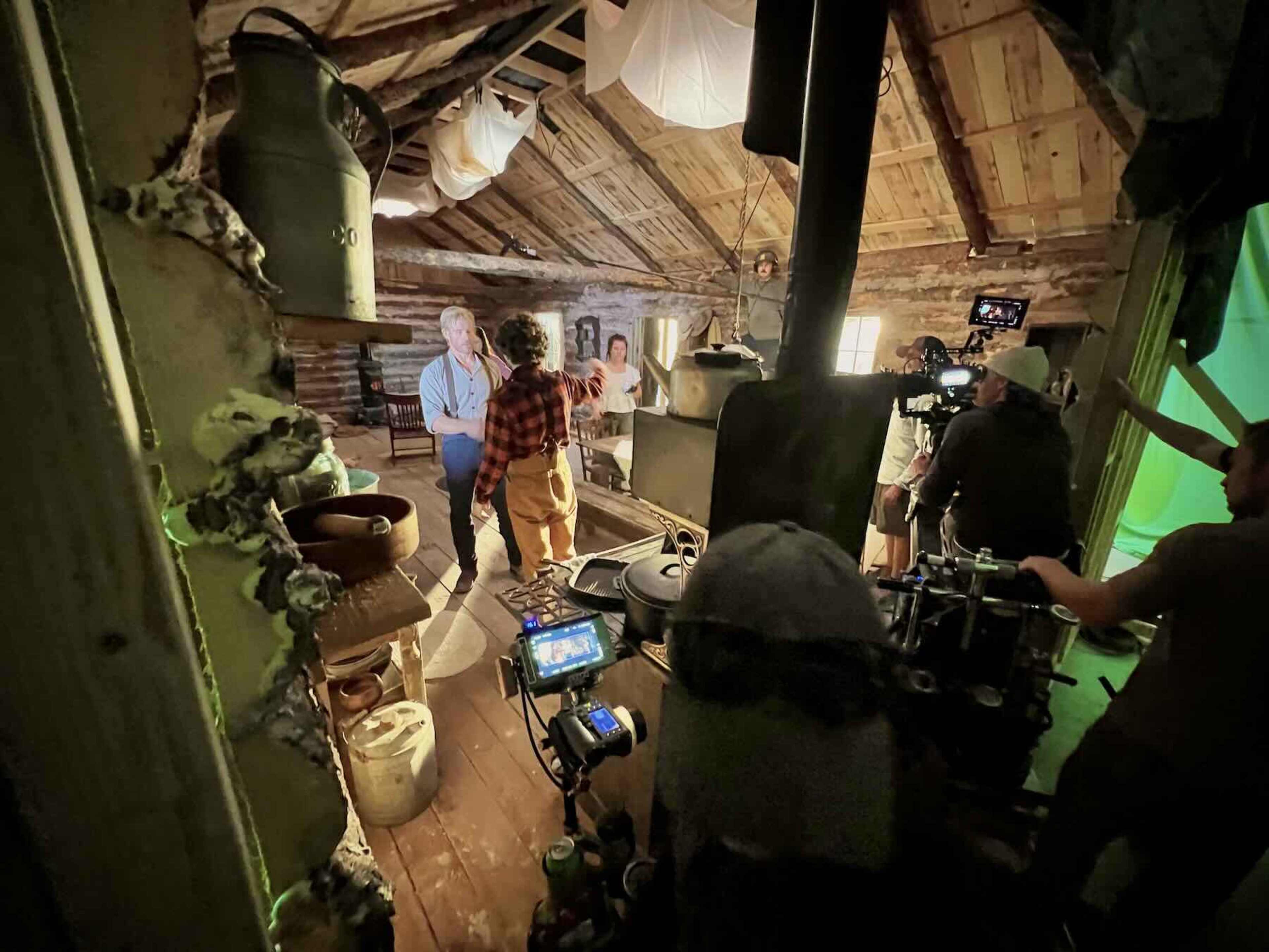 A film crew sets up a scene inside a rustic cabin with wooden beams, featuring actors in period costumes and various filming equipment.