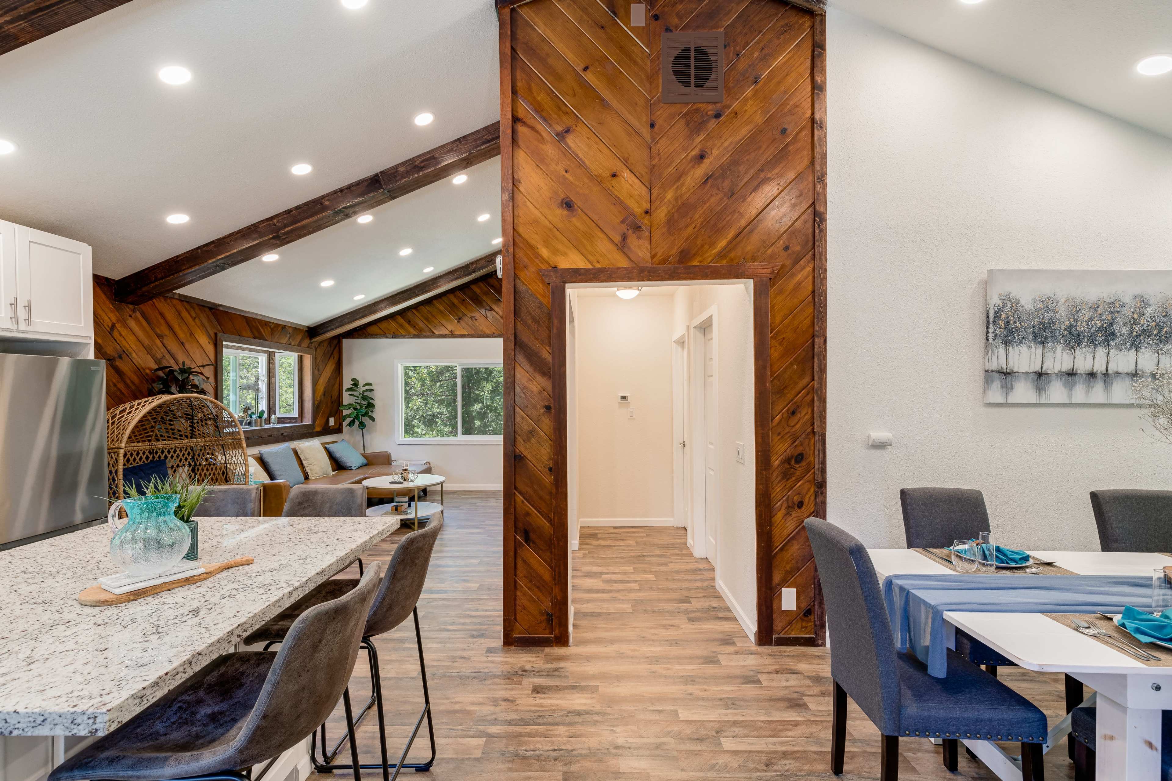 A modern interior space featuring a kitchen with a dining area on the left, a hallway leading to other rooms, and a living area with wooden walls and large windows.