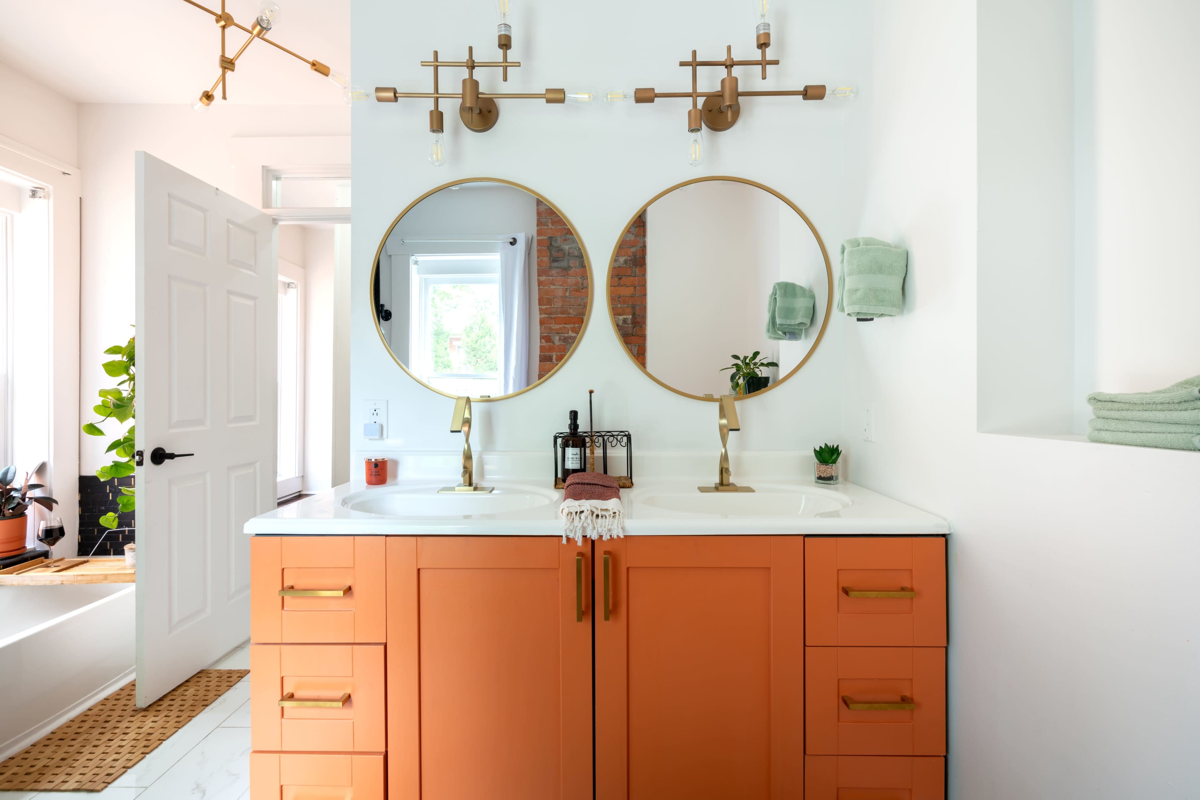 A modern bathroom with a double sink vanity featuring orange cabinets and round mirrors, complemented by wall-mounted lamps and green towels.