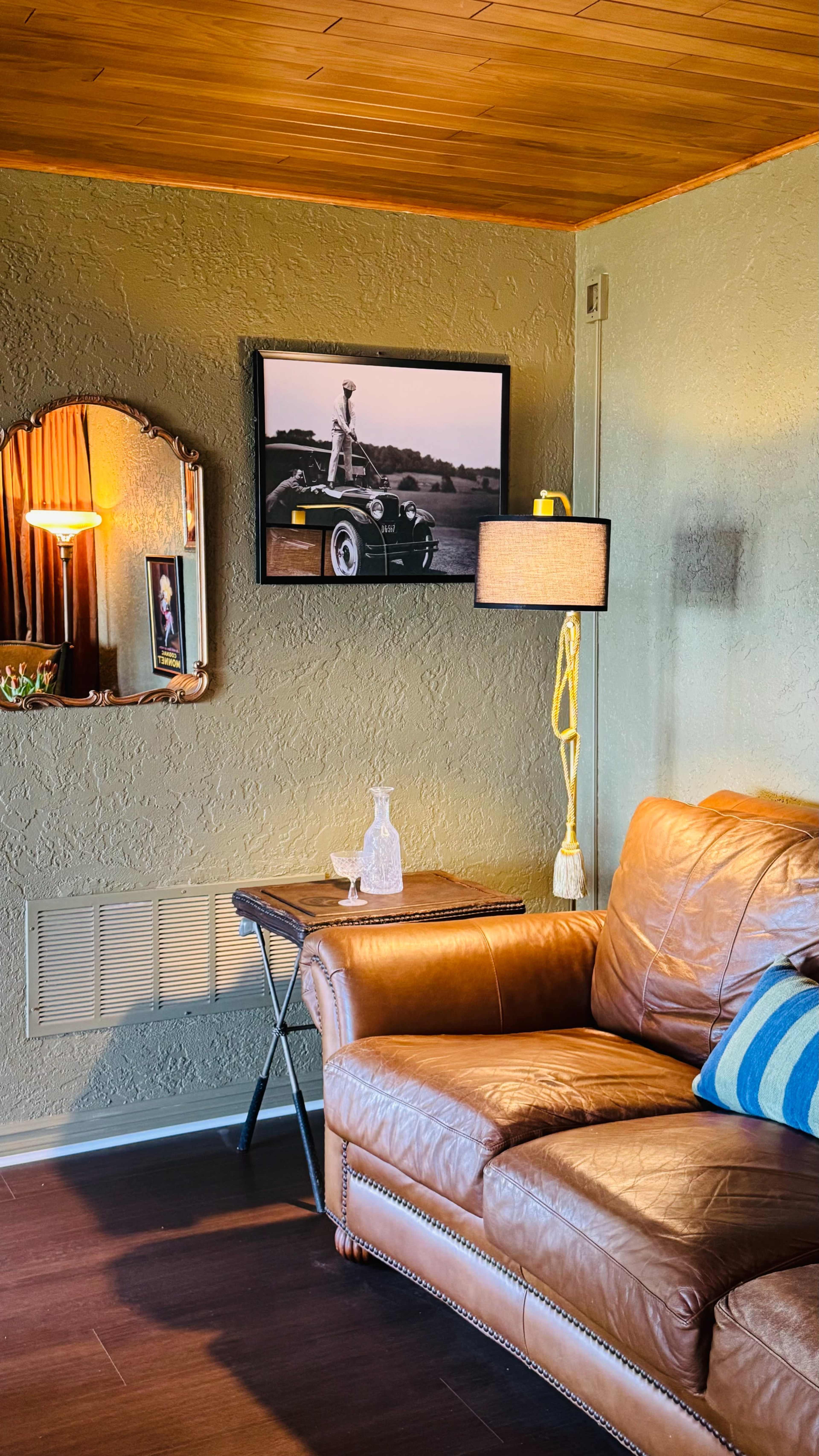 The image shows a cozy living room corner with a brown leather sofa, a framed black-and-white photo of an old car on the wall, and a small table with a decorative glass piece and a lamp.