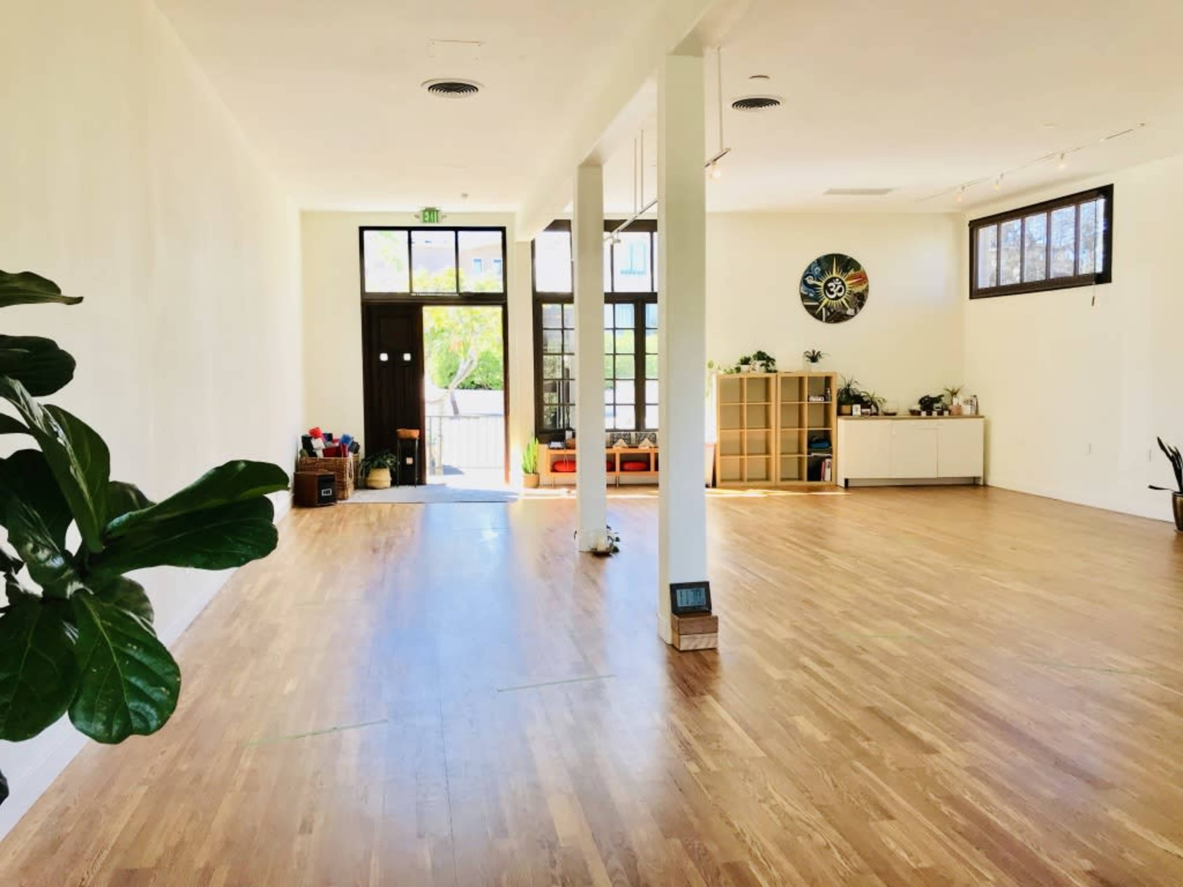 The image shows an empty room with wooden flooring, large windows, and a seating area on one side, leading to an entrance.