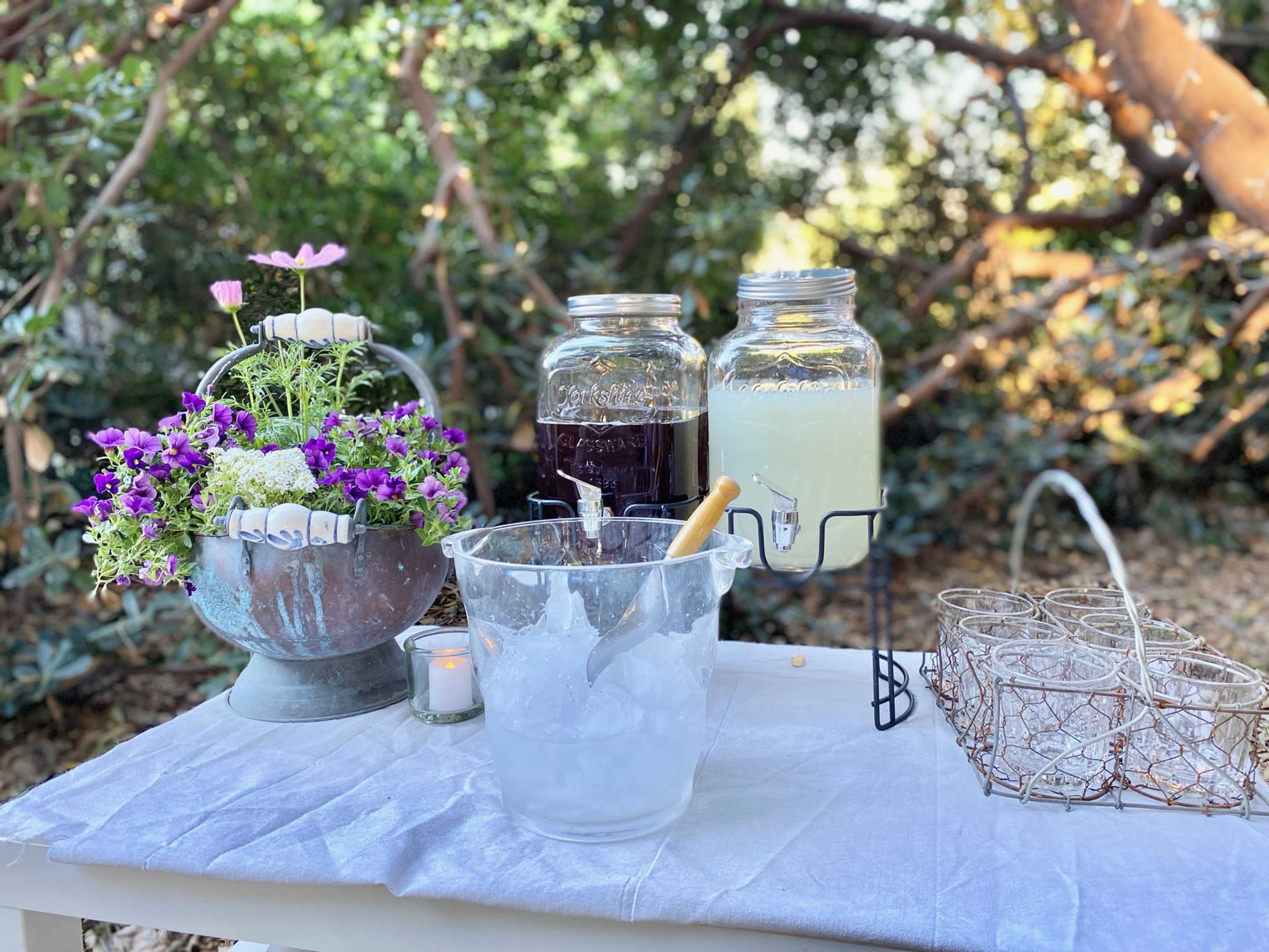 A table set outdoors with two glass jars containing beverages, a bowl of ice, a bouquet of flowers, and several glass cups.
