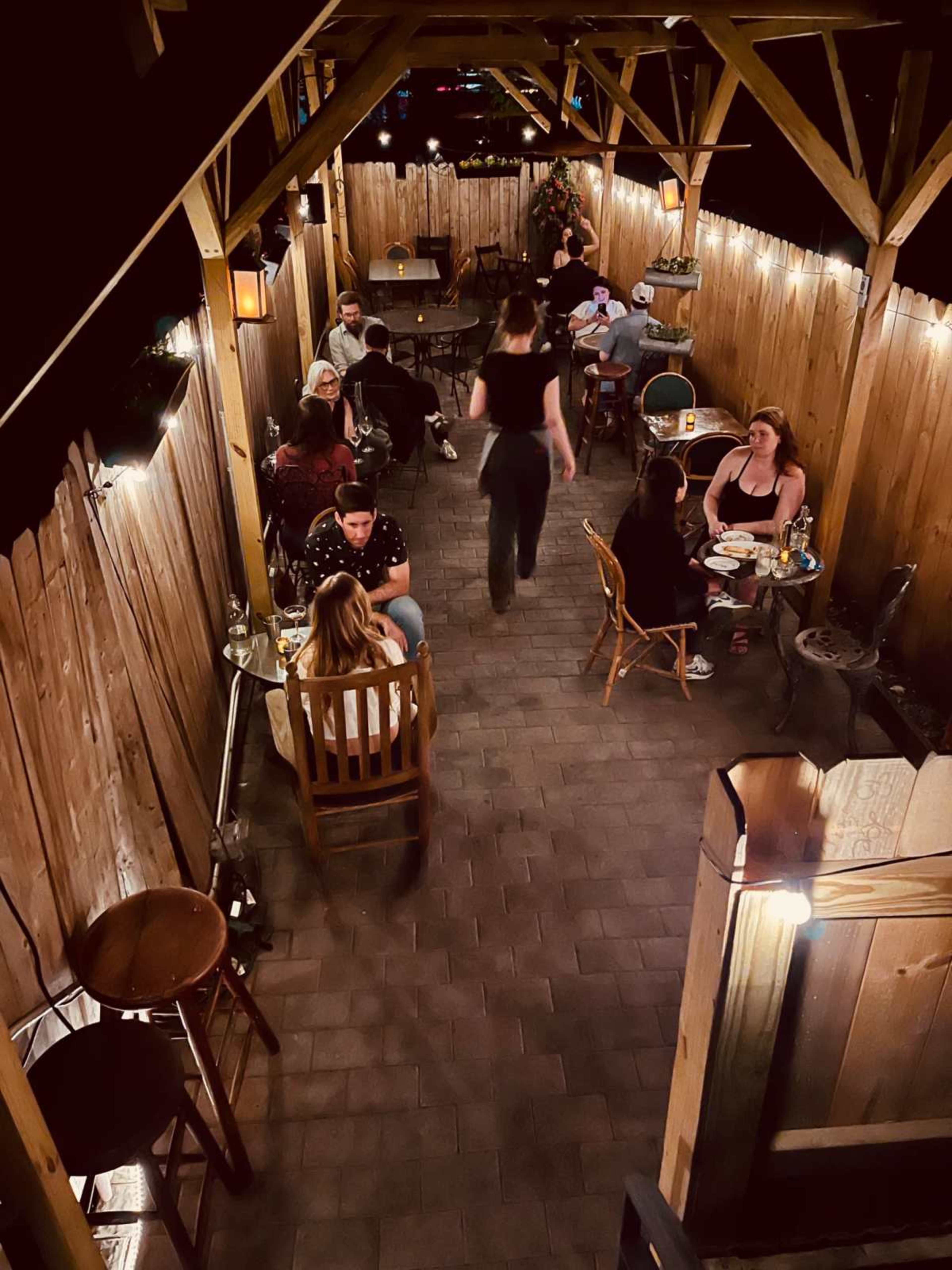 The image shows an outdoor dining area with wooden seating, illuminated by string lights, where patrons are gathered around tables.