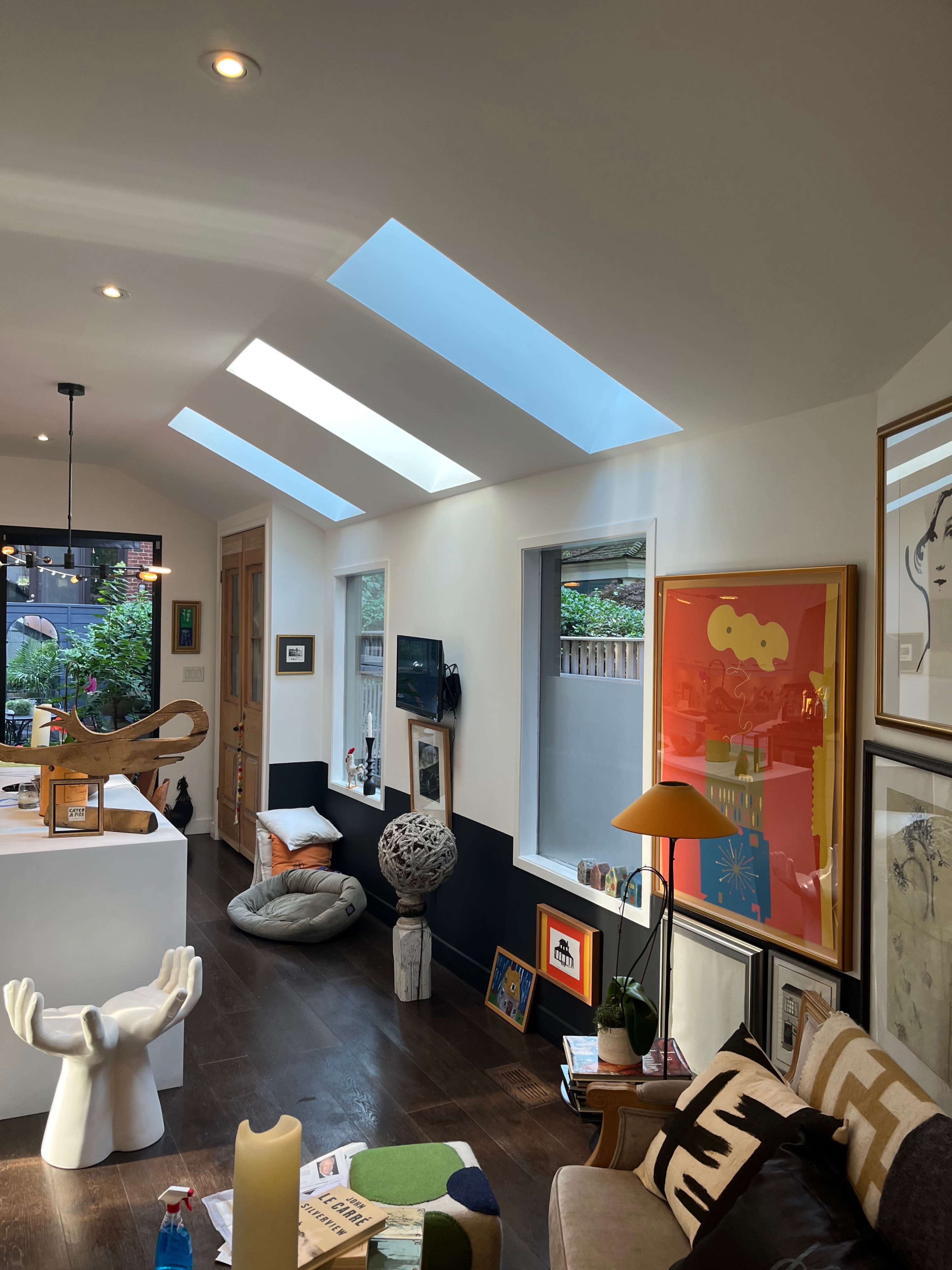 The image shows a modern interior space featuring skylights, various artworks on the walls, and a mix of decorative furniture.