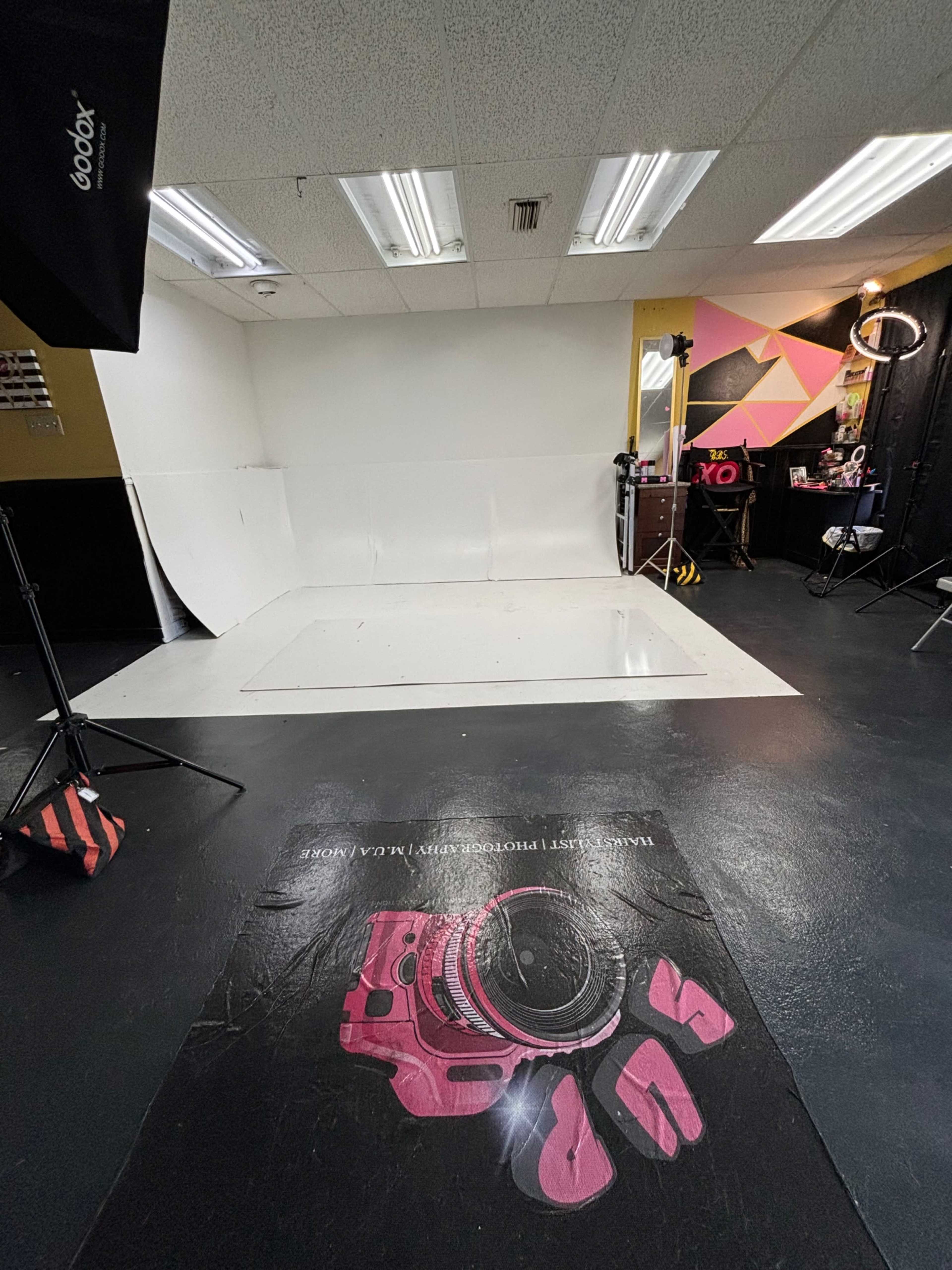The image shows a photography studio with a blank backdrop, lighting equipment, and a camera graphic on the floor.