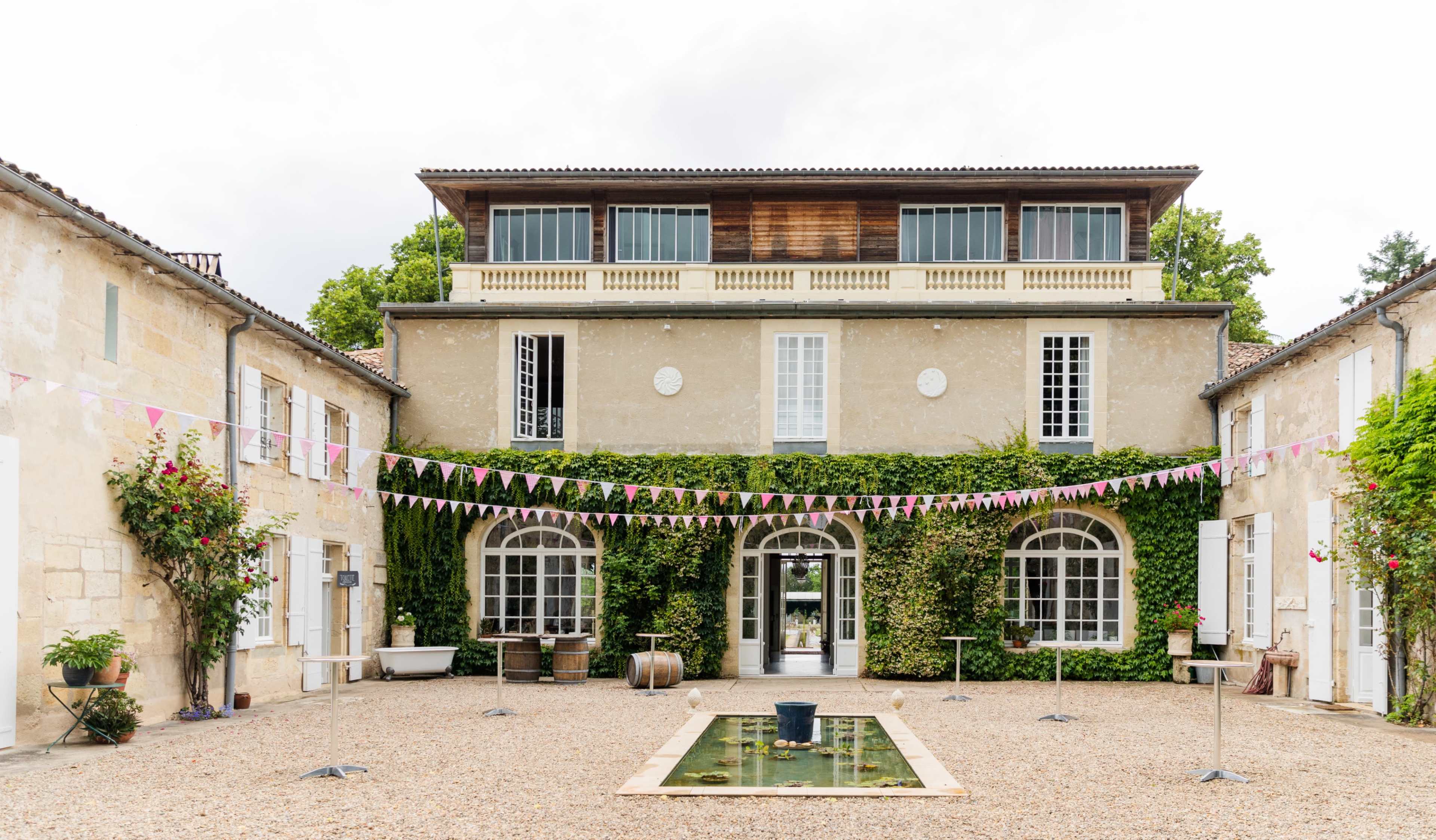 The image shows a courtyard of a stone building with arched doors and windows, surrounded by greenery and decorated with colorful banners.