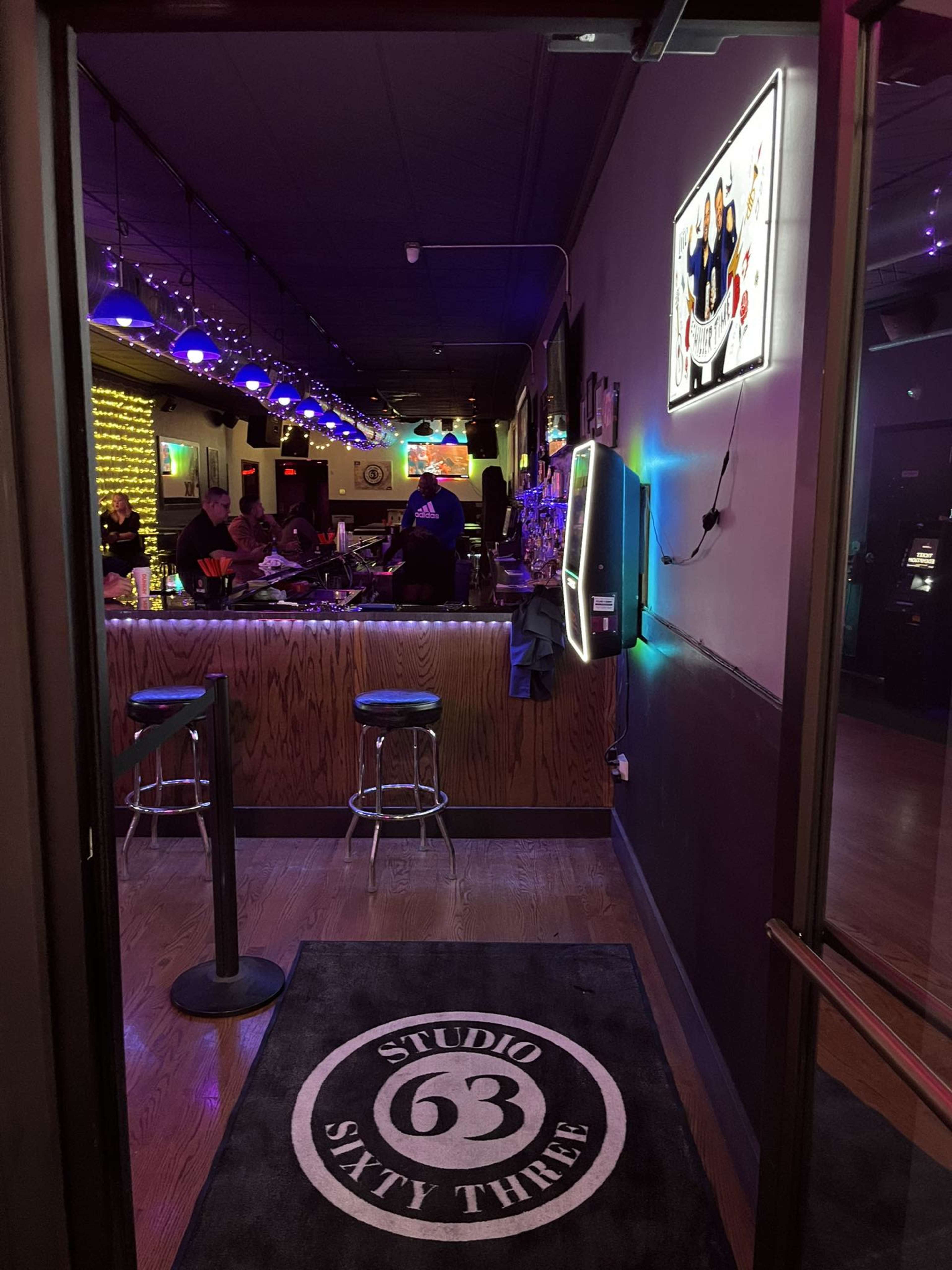 The entrance to a bar named "Studio Sixty Three," featuring a wooden bar, stools, and ambient lighting with blue accents.