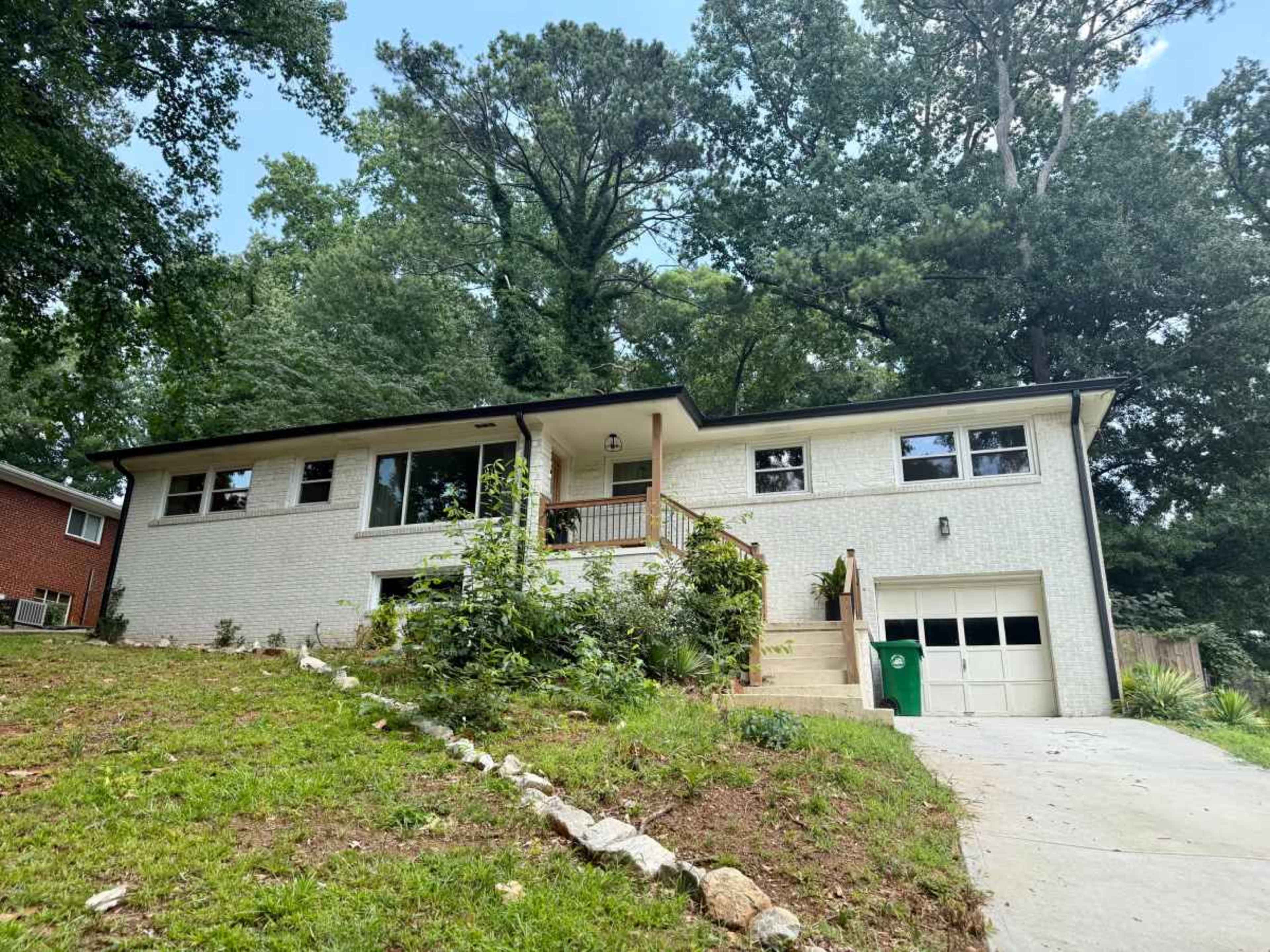 A single-story house with a brick exterior sits on a sloped lawn, surrounded by trees, with a garage and a set of stairs leading up to the front door.