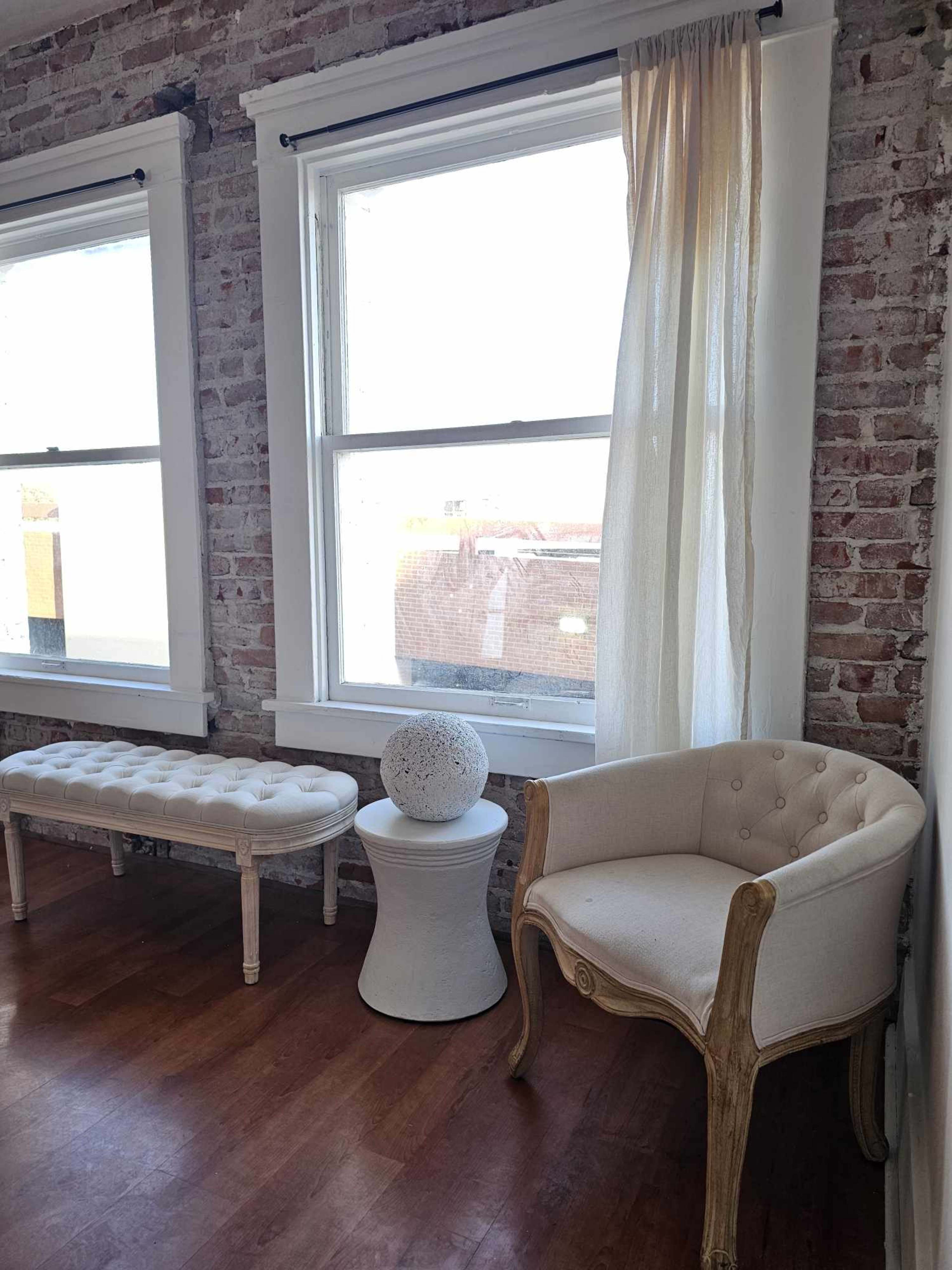 A room with a white tufted bench, a round decorative object on a white pedestal table, and an elegant armchair beside large windows with sheer curtains, against a backdrop of exposed brick walls.