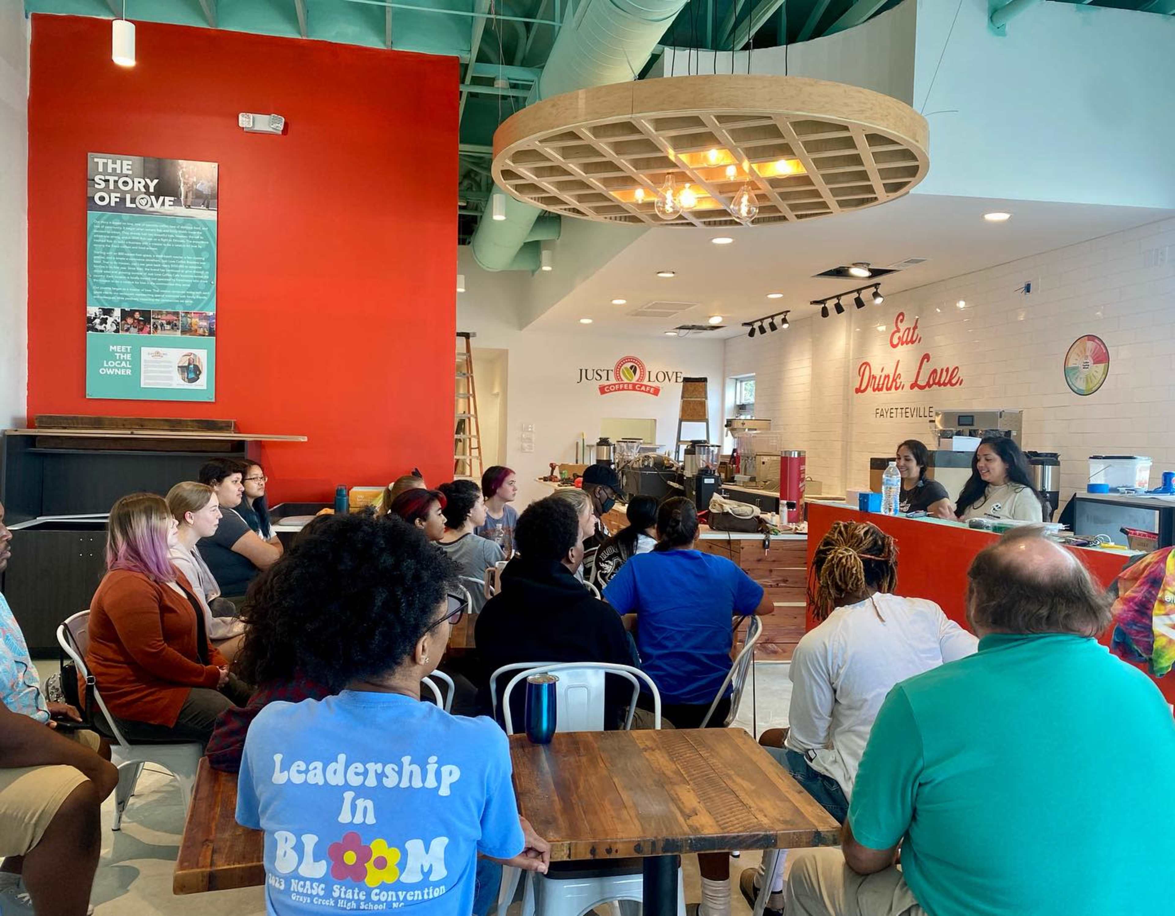 A diverse group of people is seated in a cafe, attentively listening to two speakers at the counter.