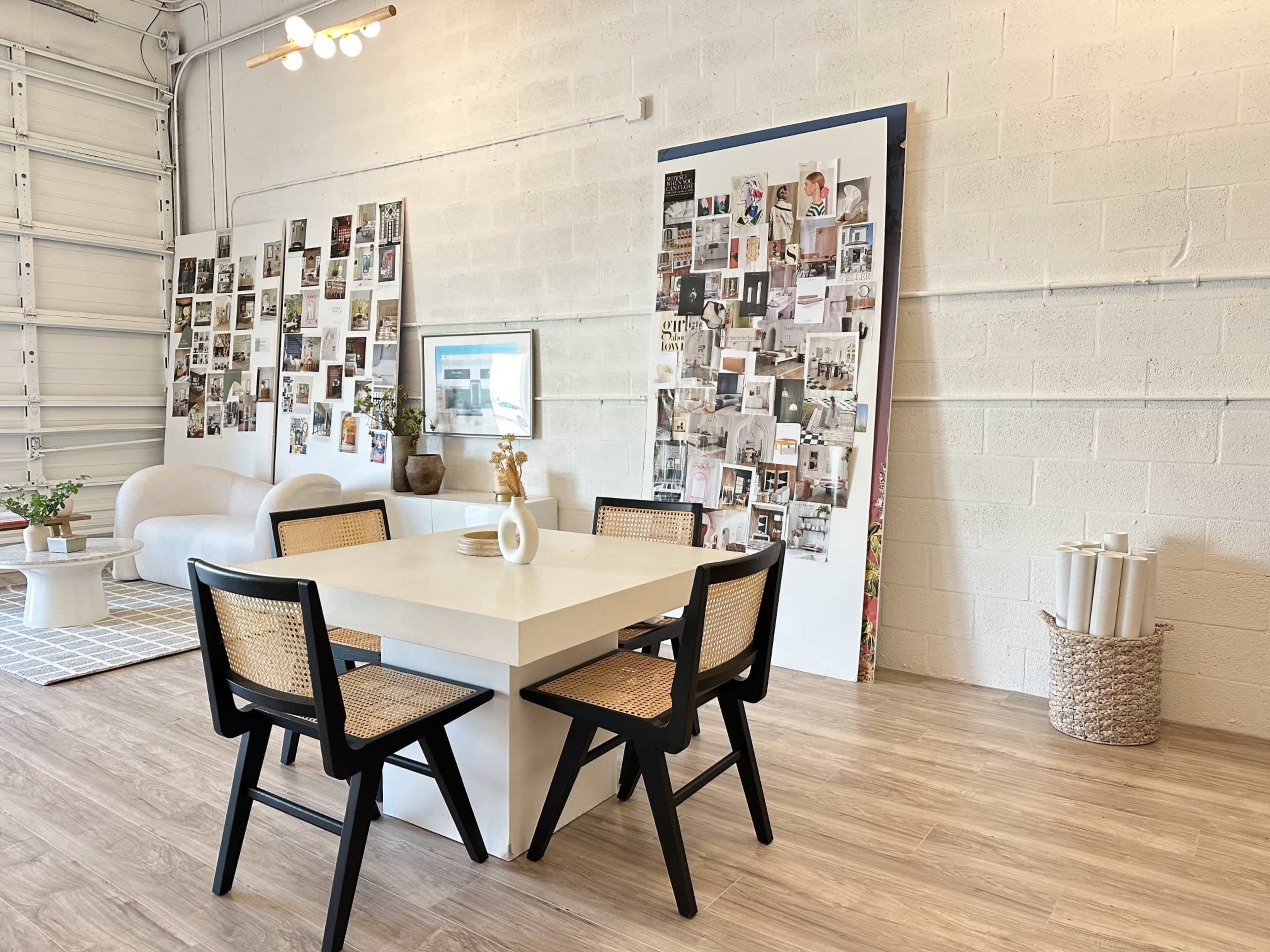 A modern dining area features a square table with four black chairs, surrounded by a gallery wall of photos and a white sofa in a spacious, light-filled room.
