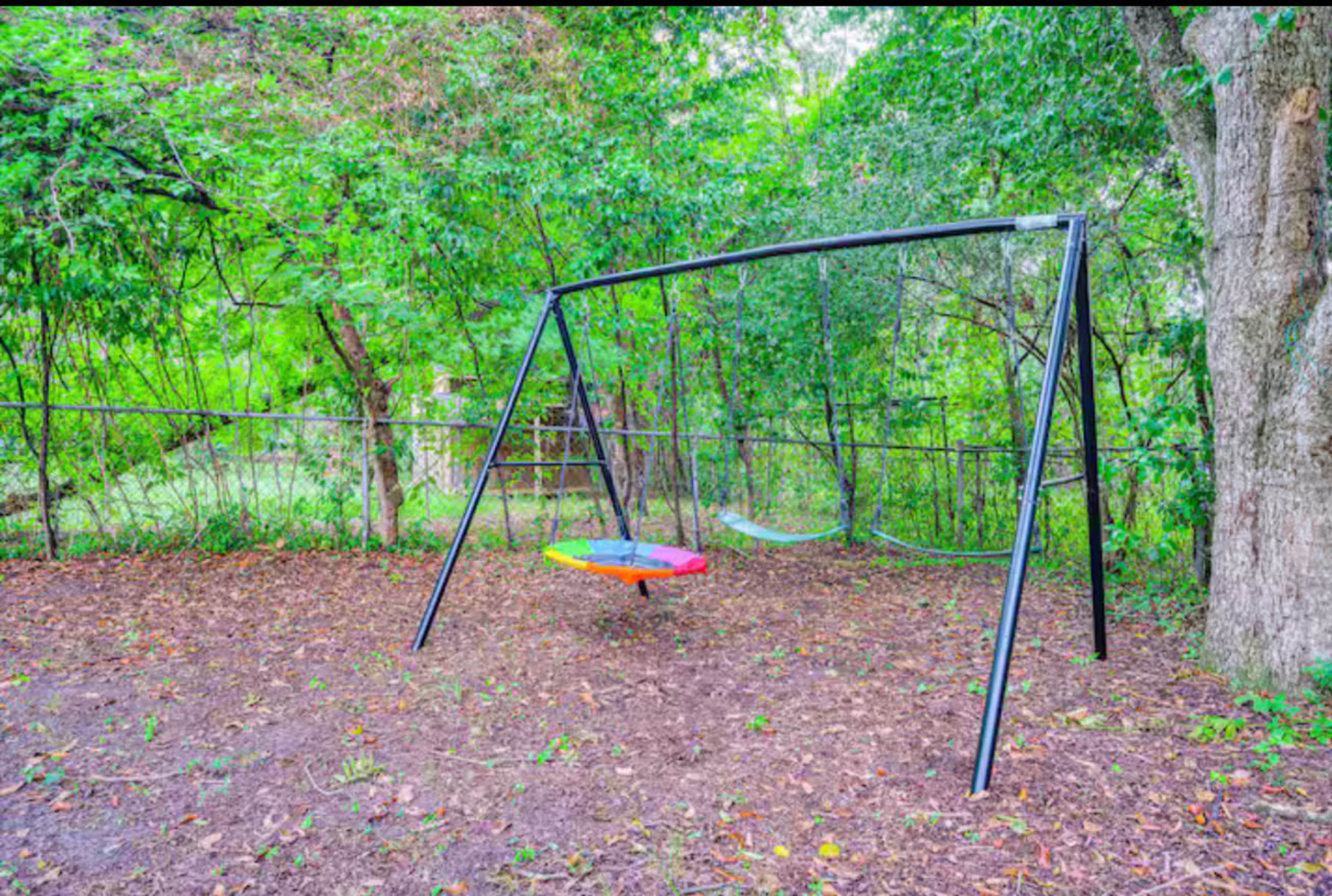 A colorful swing set with a circular swing and a traditional seat is located in a wooded backyard surrounded by trees.