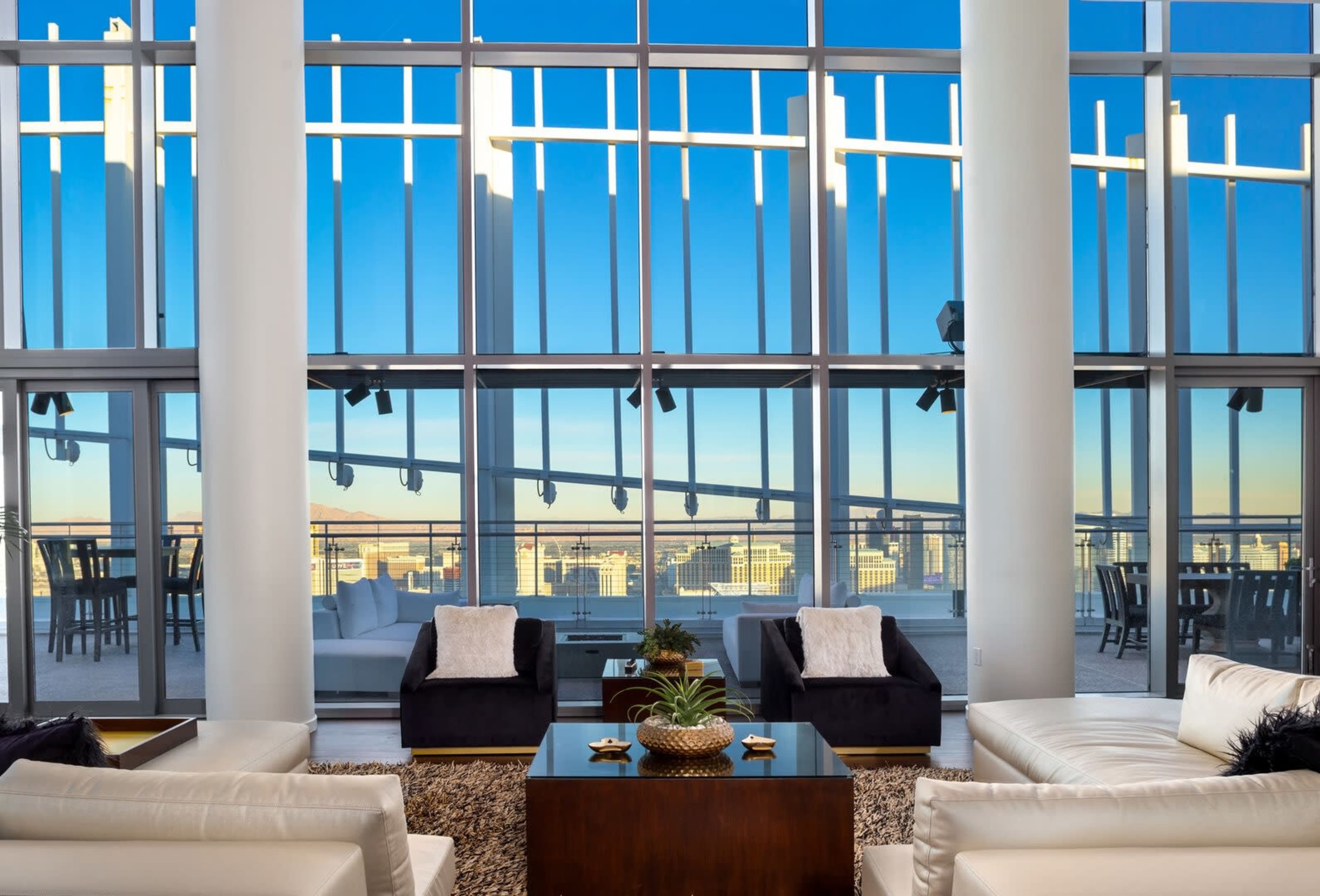 A modern living area features two contrasting sofas and a coffee table, with large glass windows showcasing a clear blue sky and cityscape in the background.