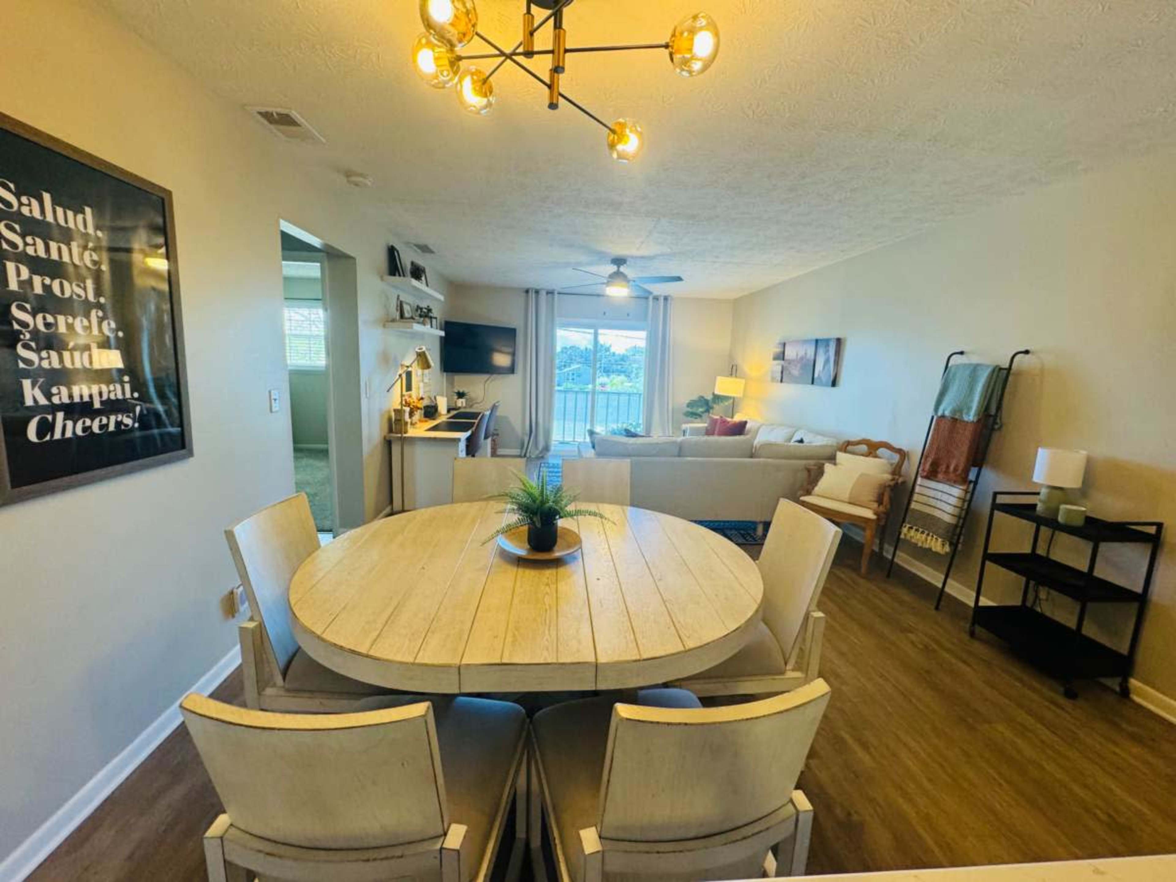 The image shows a living and dining area featuring a round wooden table with chairs, a couch, a television, and large windows allowing natural light.