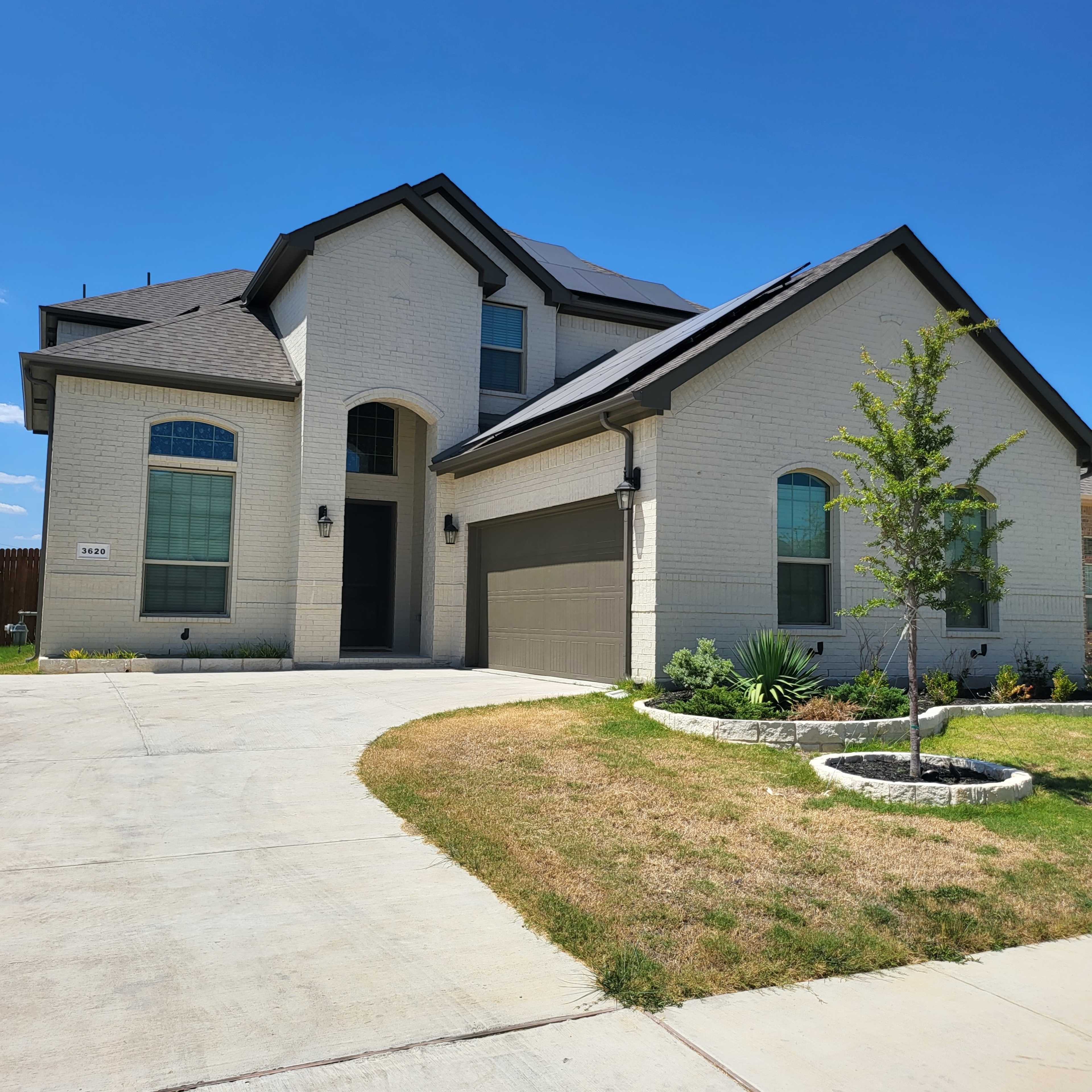 A two-story, modern-style house with a grey brick exterior and solar panels on the roof sits on a landscaped lot with a concrete driveway.