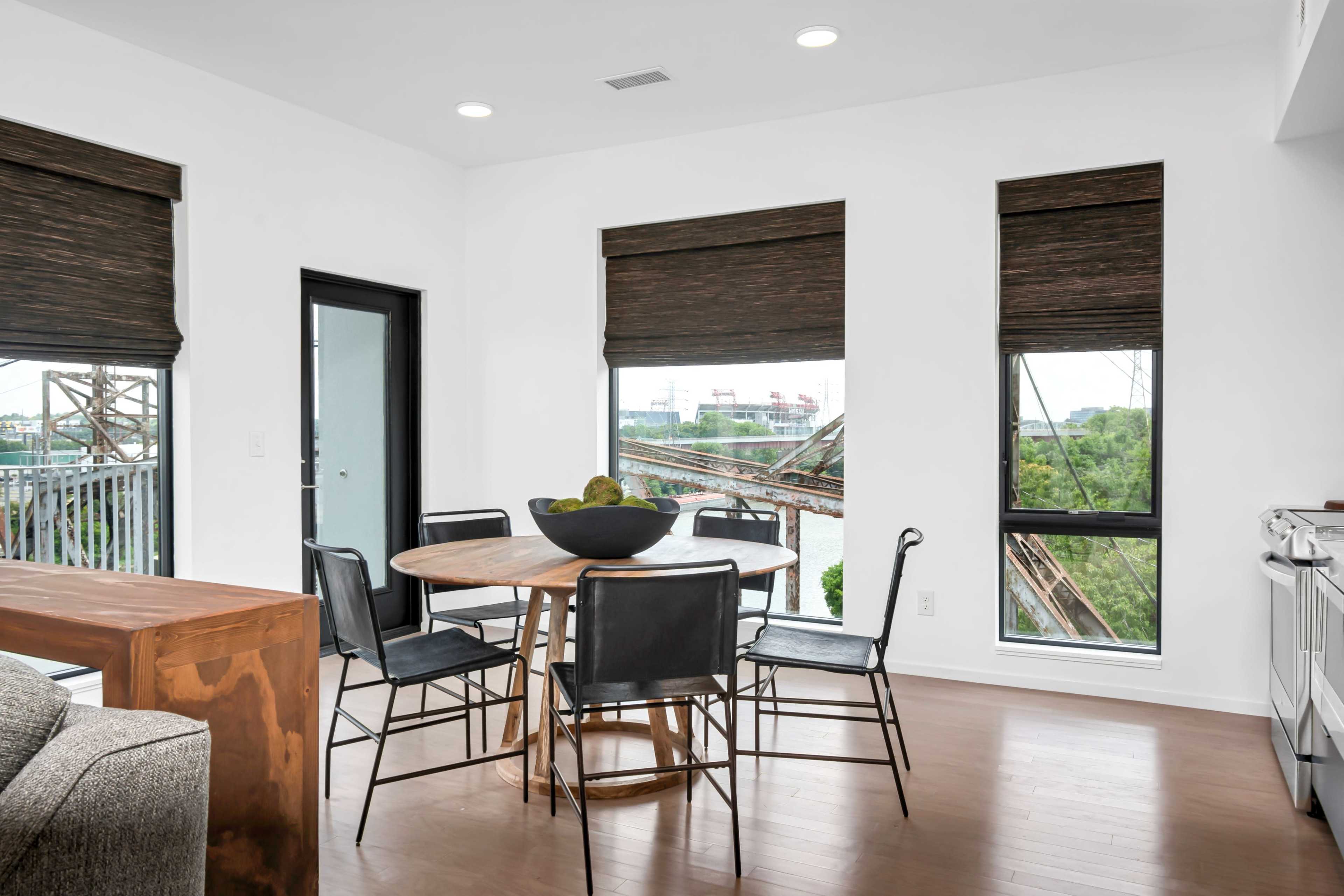 A modern dining area with a round table surrounded by chairs, large windows with shades, and a view of an industrial landscape outside.
